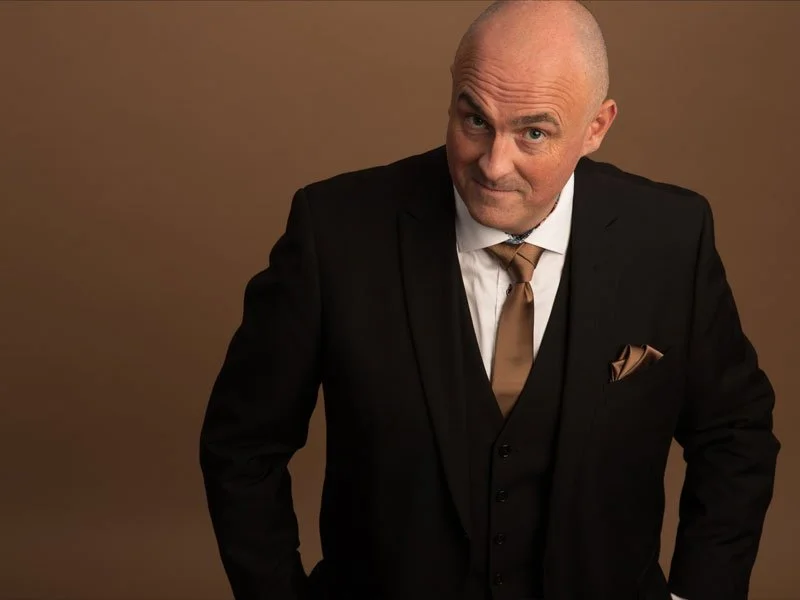 A bald man in a black suit with a white shirt and brown tie, standing against a brown background.