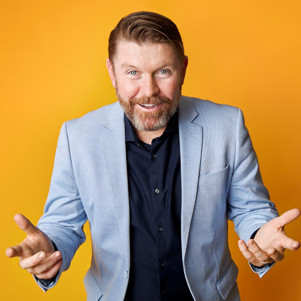 Man with a beard smiling, wearing a light blue blazer and dark shirt, gesturing with open hands against a yellow background.