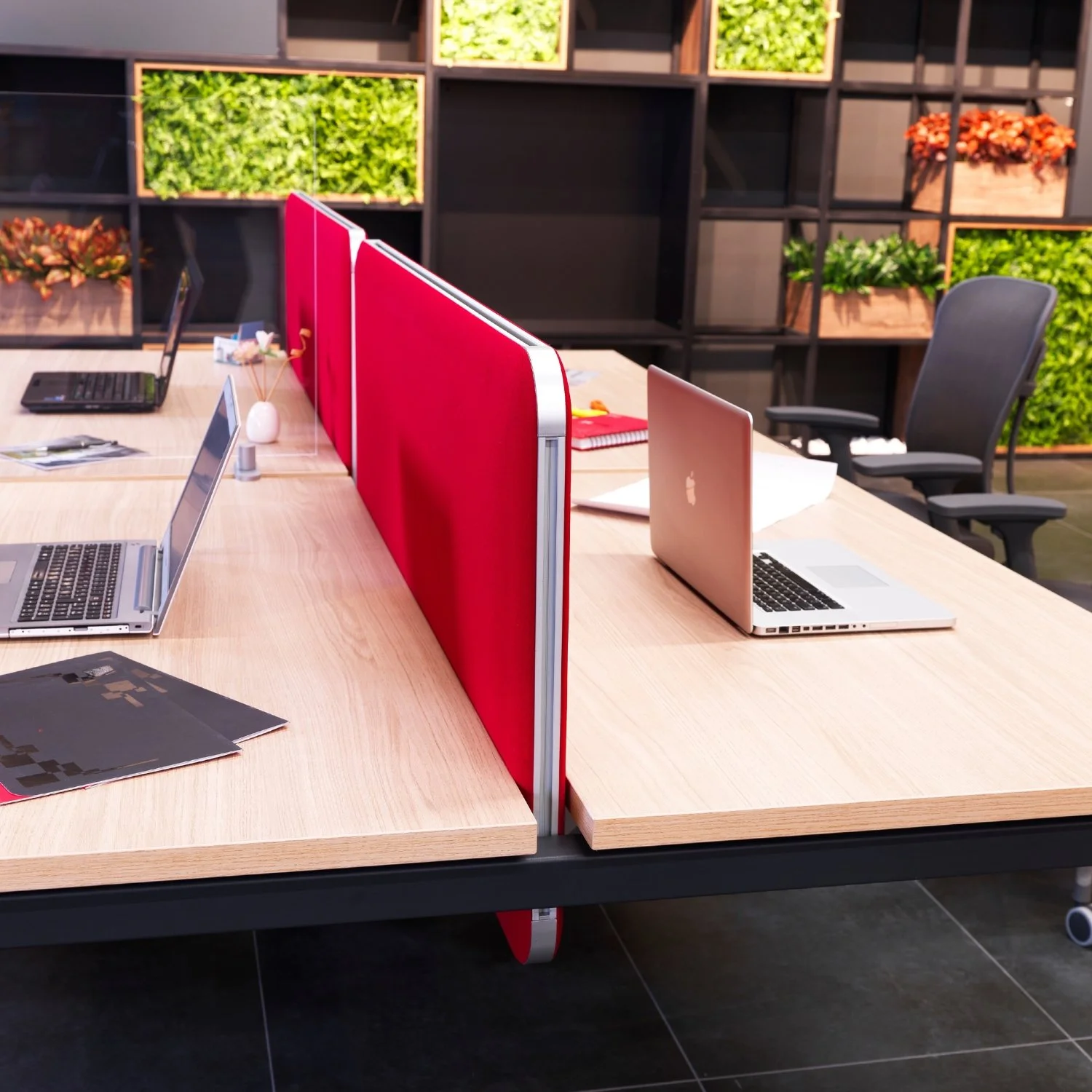 Office workspace with two desks divided by red fabric partitions, equipped with laptops, notebooks, and office supplies, set against a wall with green and orange plants.