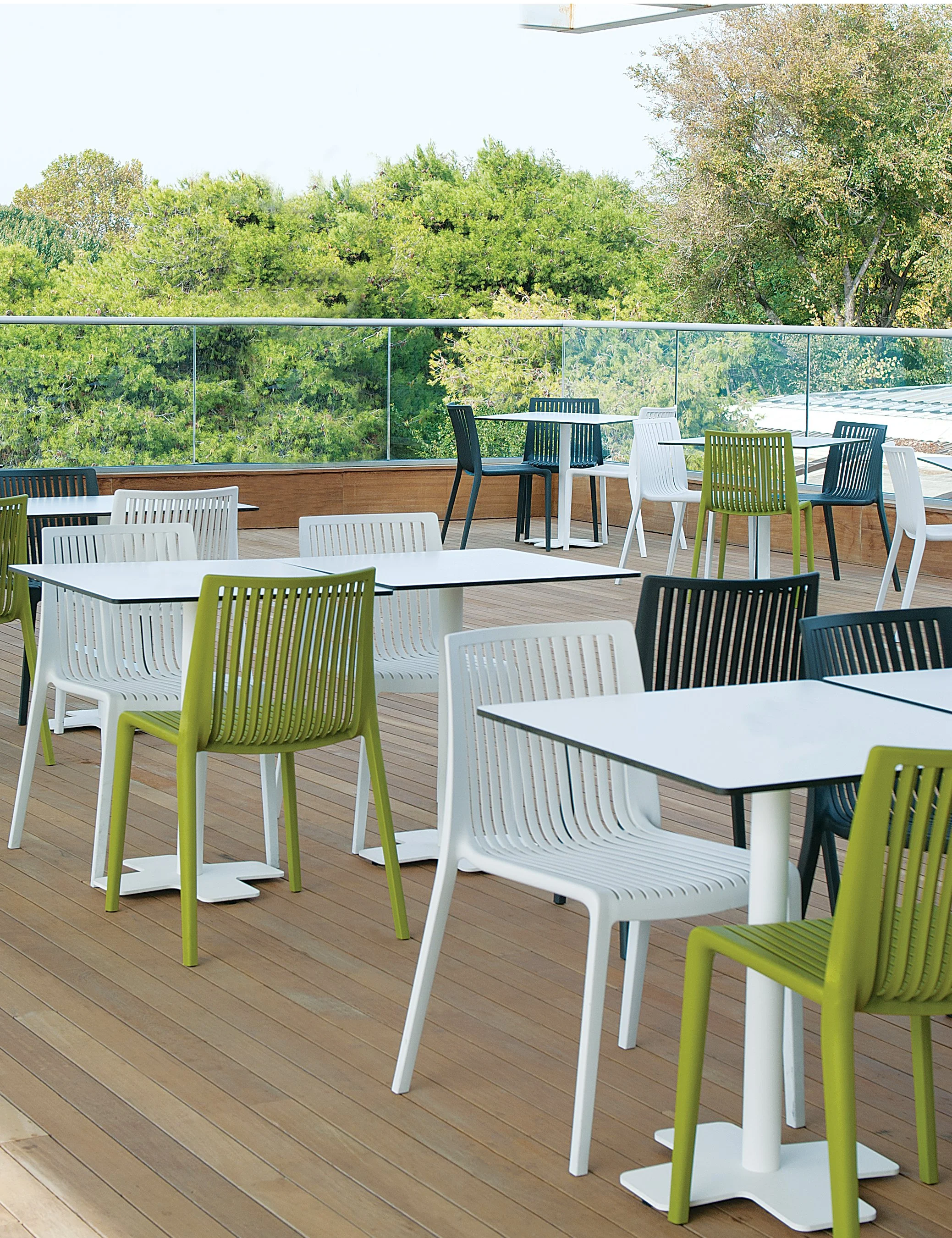 Outdoor seating area with white, black, and green chairs and tables on a wooden deck, with a glass railing and trees in the background.