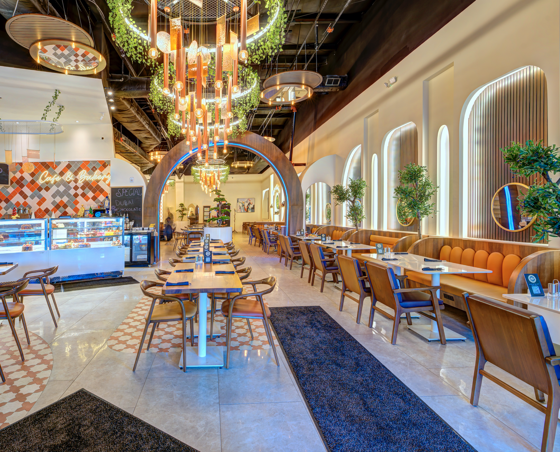 Interior of a modern restaurant with a high ceiling, decorative lighting fixtures, and large arched windows. There are wooden chairs and tables arranged neatly, with a cozy seating area along the wall decorated with green potted plants. A counter with a display case of desserts and a chalkboard sign is visible in the background.