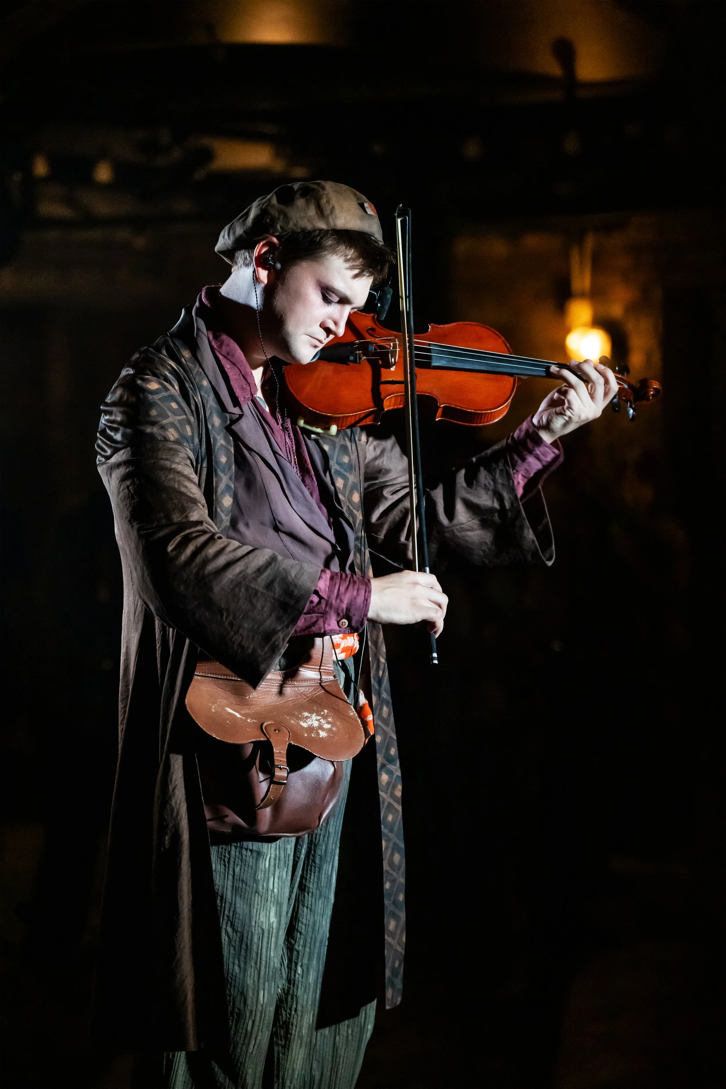 A young man playing the violin on stage, wearing a hat, patterned coat, and striped pants, with a brown leather shoulder bag and a dark background.
