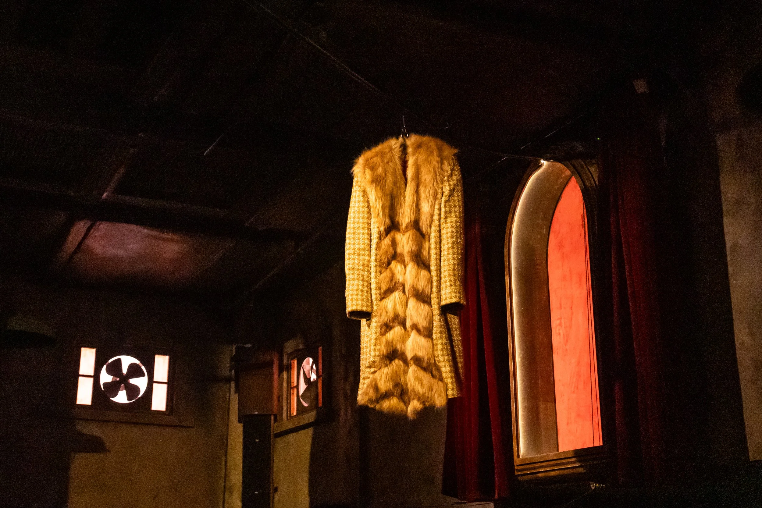 A vintage coat with a fur collar and fur accents hanging on a wall next to a curved, illuminated wooden frame with a red curtain behind it.