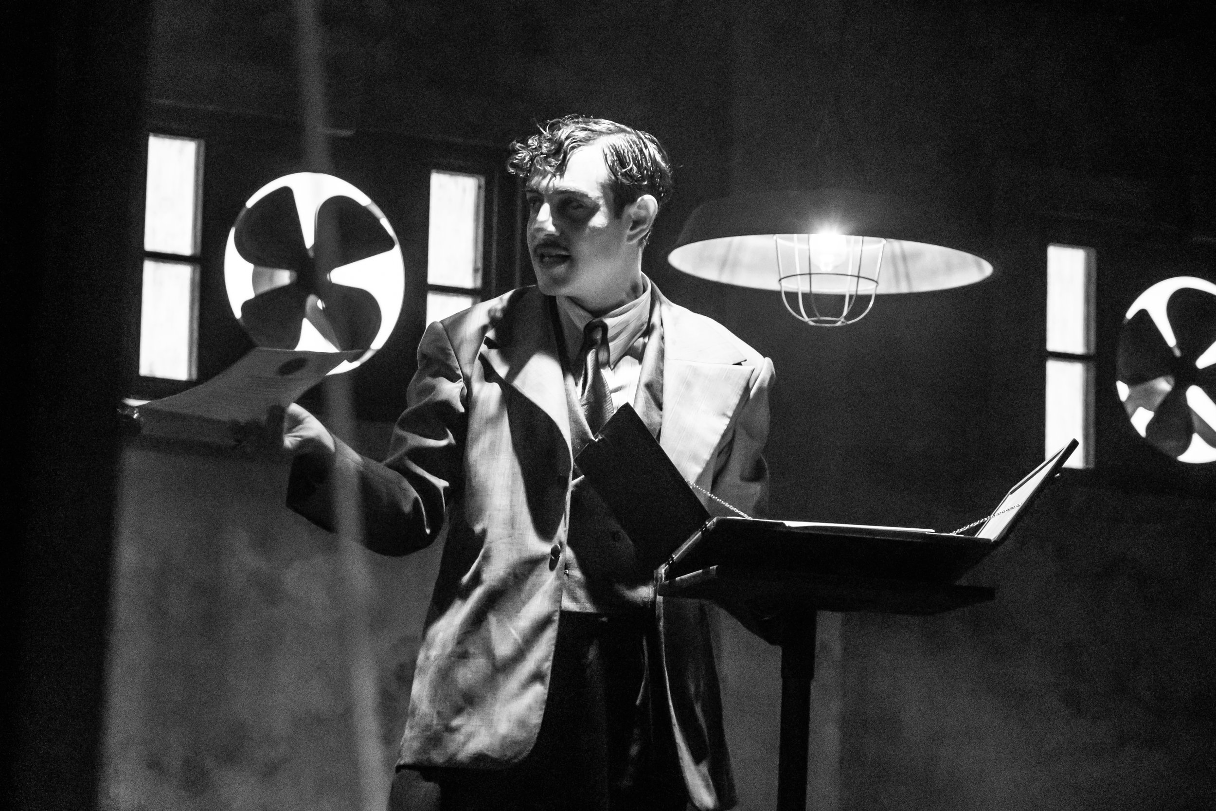 A black and white image of a man with slicked-back hair, wearing a trench coat, standing at a music stand with an open book or folder. The man appears to be singing or speaking passionately. There are large circular fans with propeller blades on the 