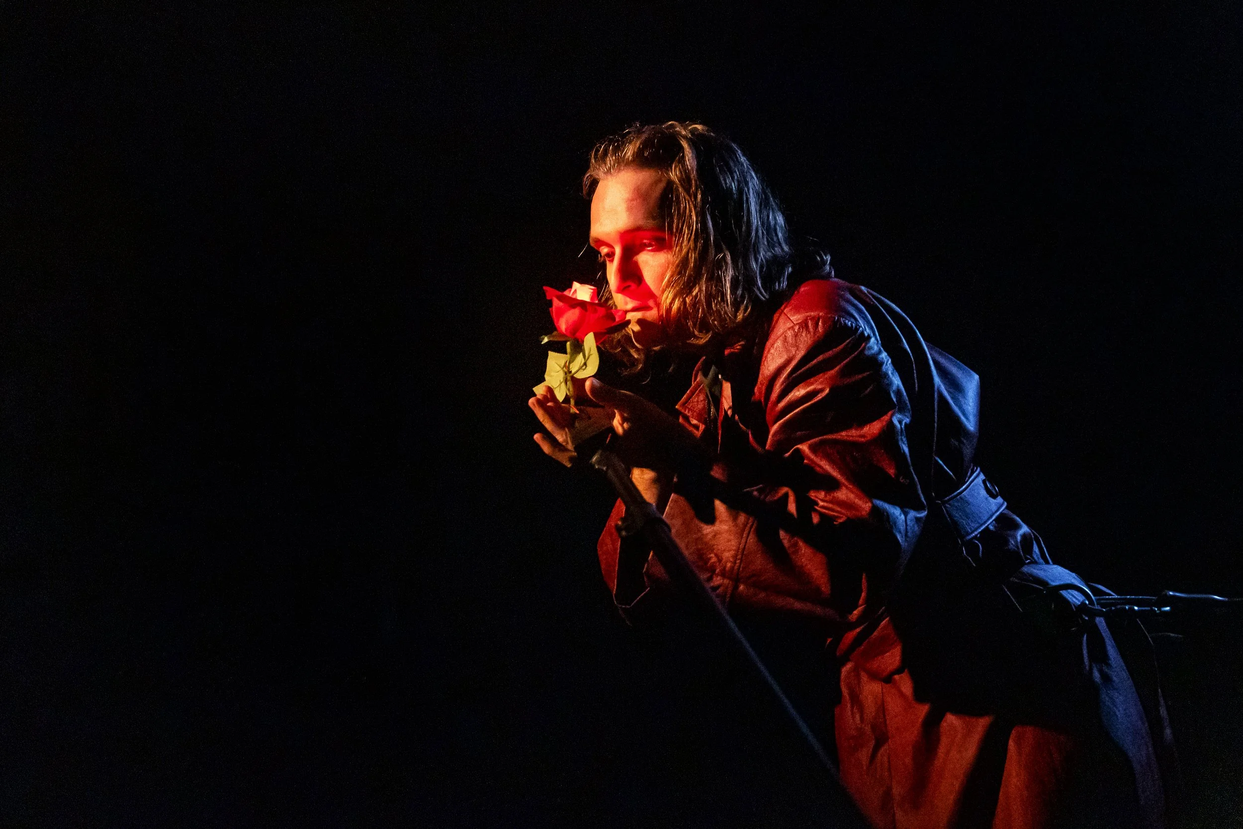 A man in a dark environment is holding a red rose close to his face, illuminated by a soft light, with it partially obscured by the darkness.