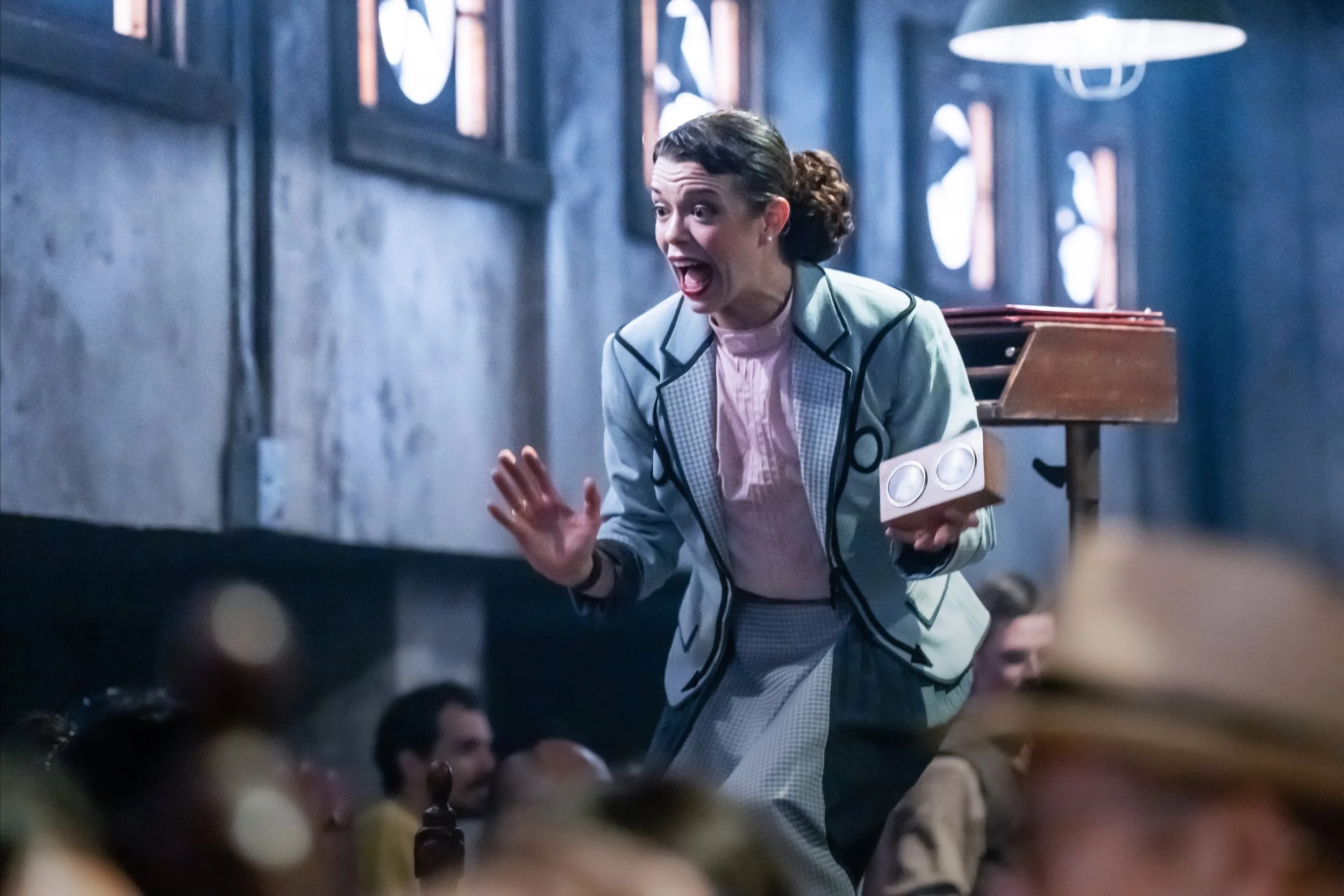 A woman in a light grey blazer and checkered skirt, holding a clipboard, gesturing to a group of people in a dimly lit room.