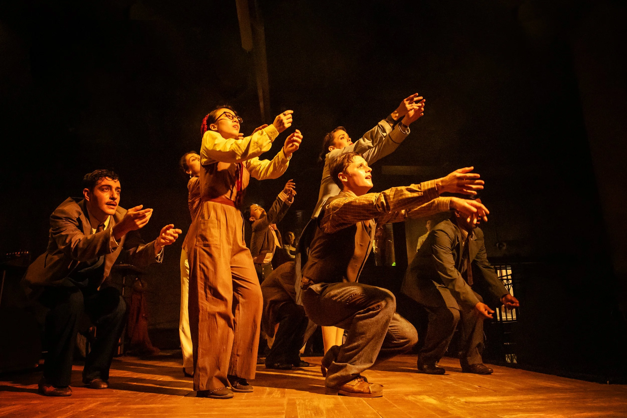 Actors performing on stage, kneeling and reaching out as part of a theatrical scene, with warm lighting emphasizing their expressions and movements.