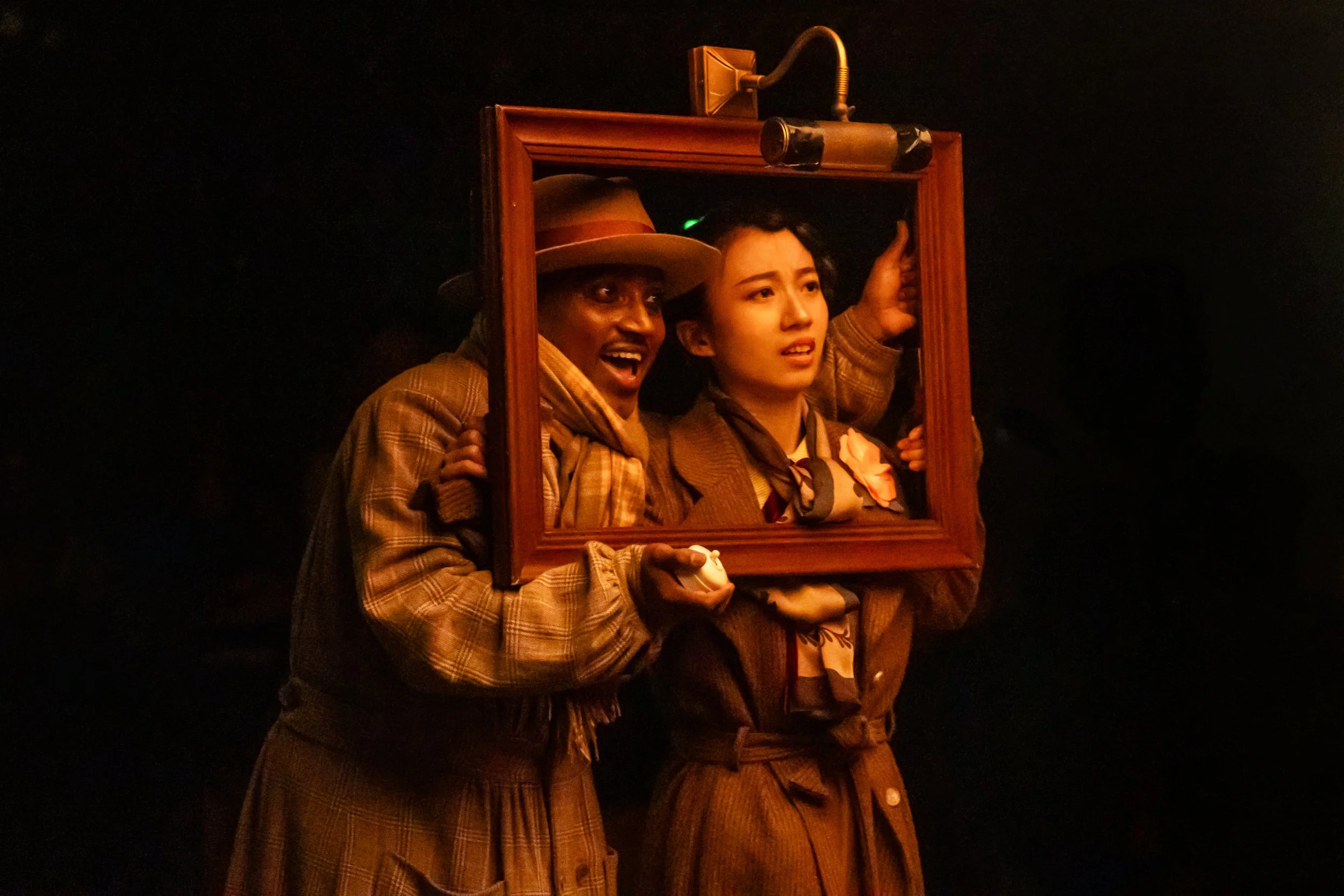 Two actors in vintage clothing holding a picture frame as a prop, with one actor appearing surprised or excited and the other looking concerned, against a dark background.