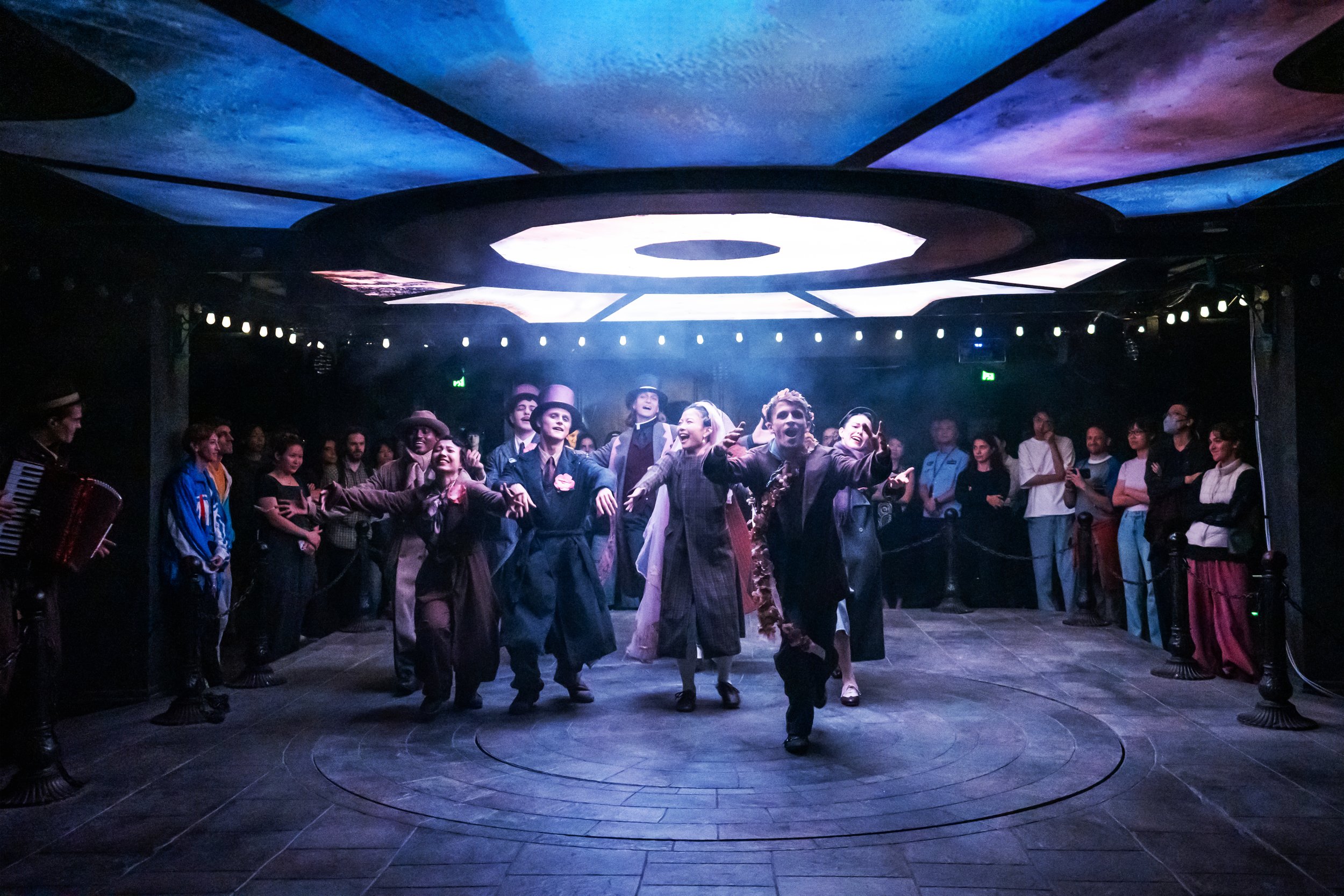 Group of performers dancing in a theatre with audience watching, stage illuminated with colourful lights.