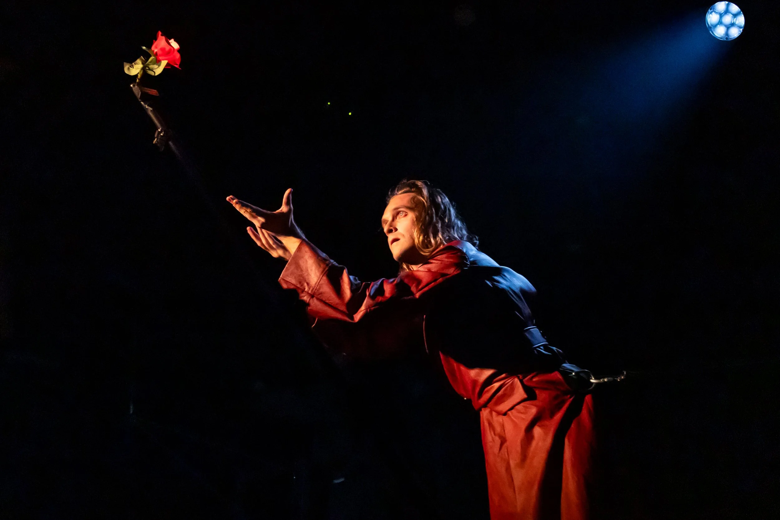 Performer in a red and black costume reaching for a red and yellow rose on a dark stage, illuminated by a spotlight.