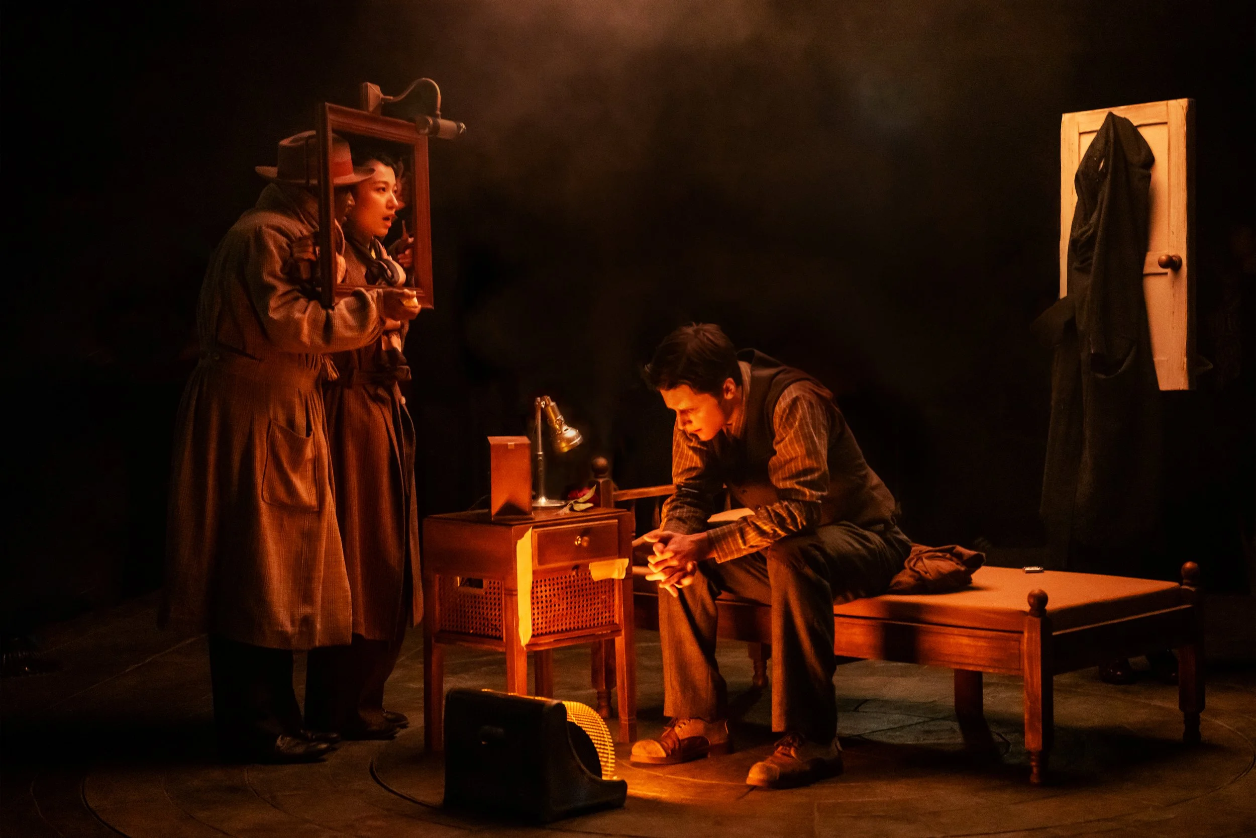 Three actors on a stage with dim lighting, one sitting on a bed and two standing nearby, in a scene resembling a play set in an indoor room.