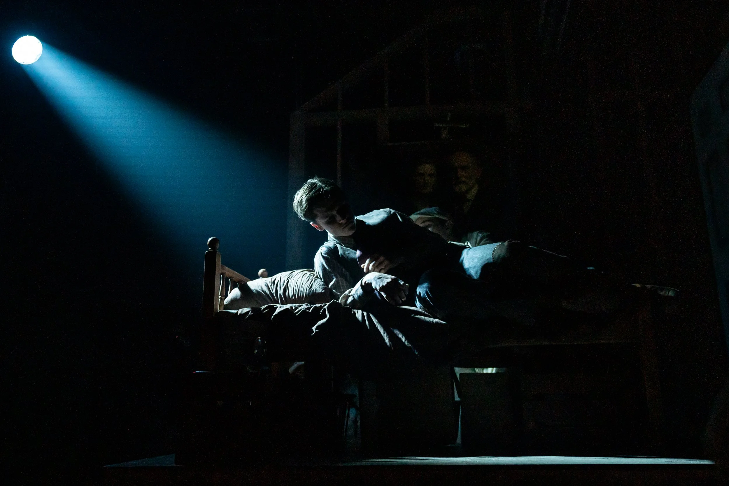 A person lying on a bed in a dark room, illuminated by a spotlight from the ceiling, with a poster of two people faintly visible in the background.