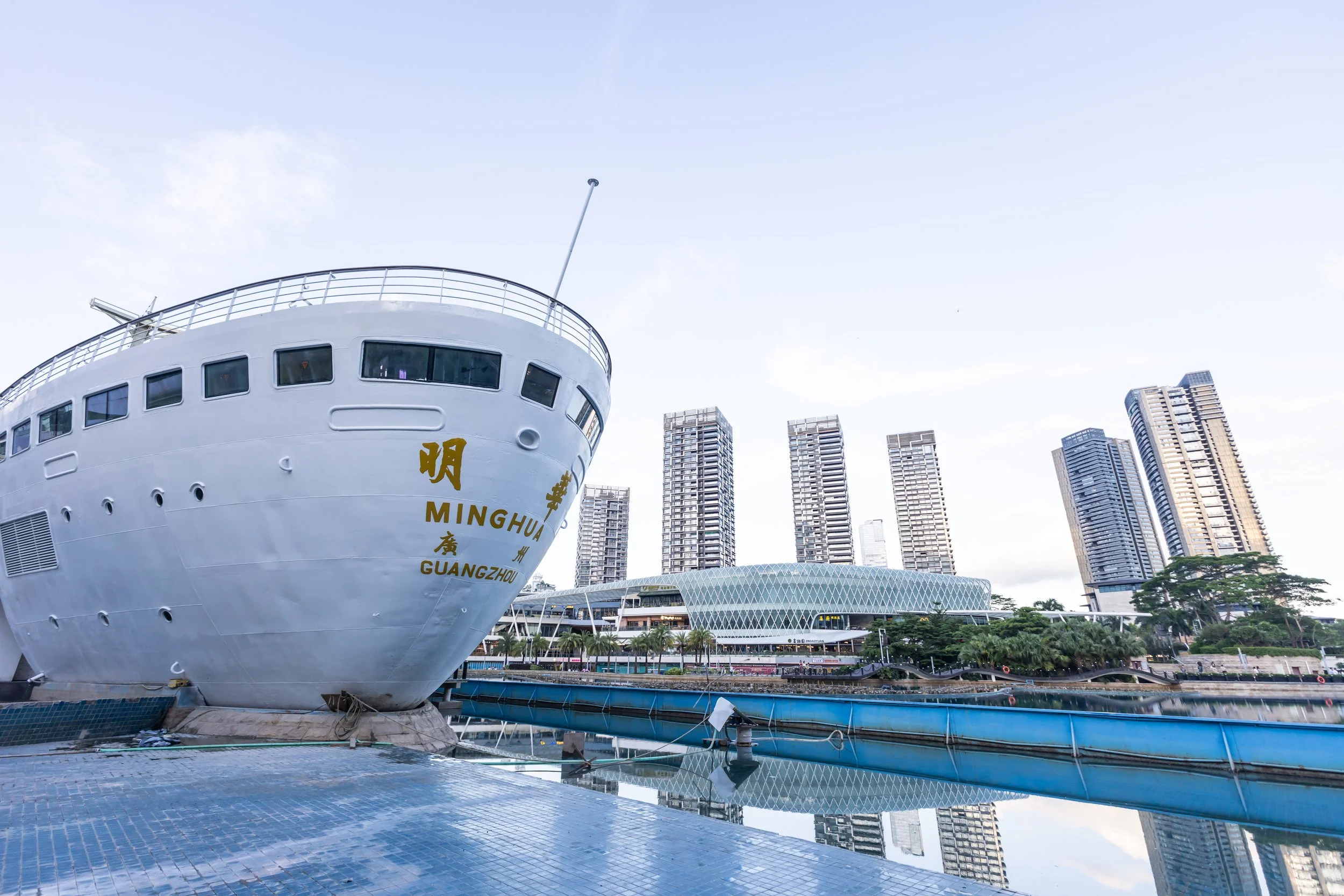 Large white ship with Chinese characters and the words MINGHUA in yellow, docked near a body of water. Modern high-rise buildings and a domed structure are visible in the background, under a partly cloudy sky.