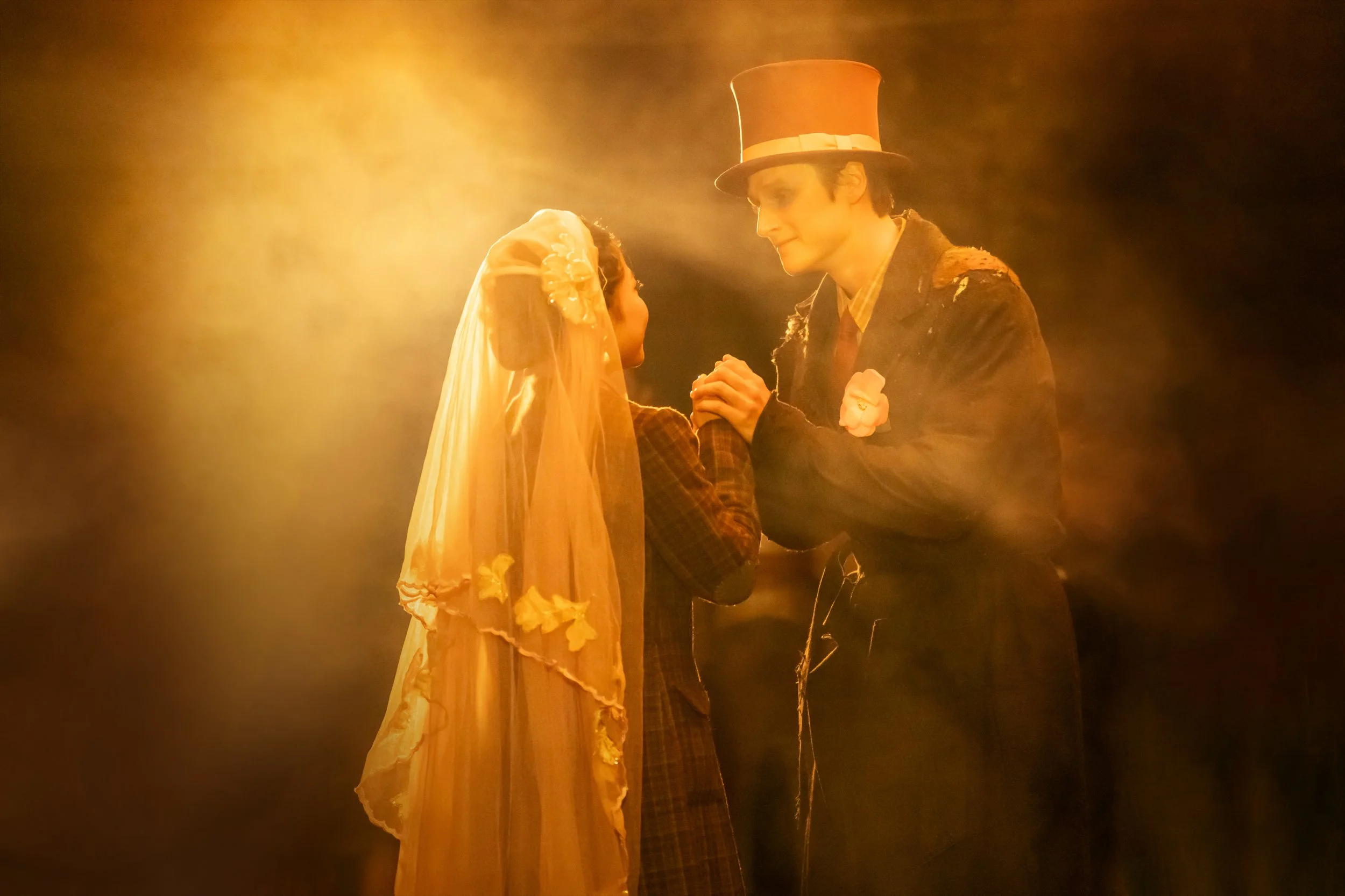 A man and a woman dressed in vintage clothing share a tender moment on stage. The woman wears a long, yellow, ruffled dress and veil, while the man wears a dark coat, a plaid shirt, and a tall top hat. They are holding hands and looking into each oth