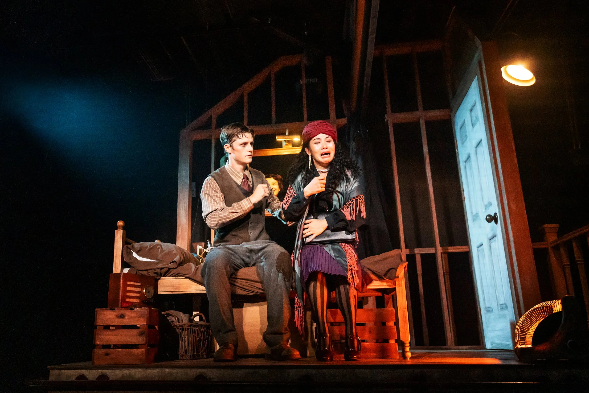 The scene shows two actors performing on stage, with one seated on a bed and the other standing. The person standing appears emotional, possibly crying, and is dressed in colorful, layered clothing with a headscarf. The stage set includes a bed, wood