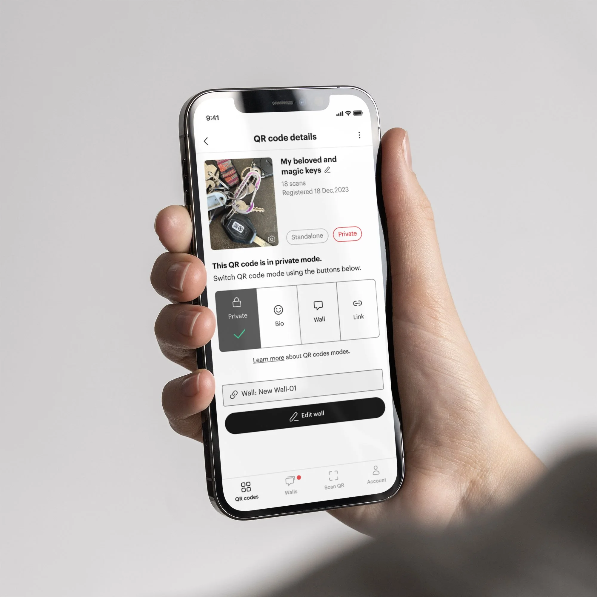 Person holding a smartphone displaying an app with QR code details. The screen shows a section titled 'QR code details' with an image of keys labeled 'My beloved and magic keys.' The mode is set to 'Private.' Options for 'Bio,' 'Wall,' and 'Link' are visible below.