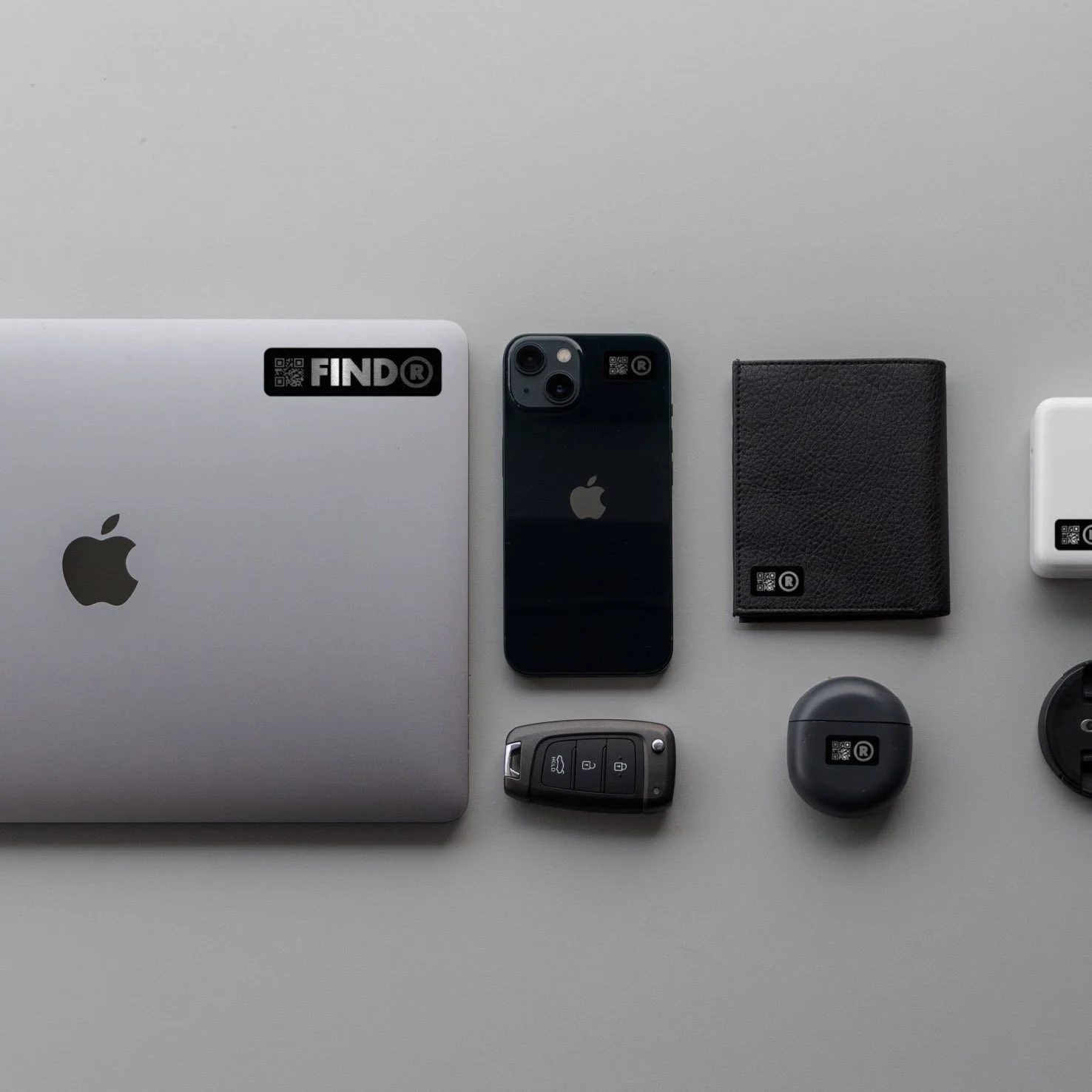 A flat lay of electronic and personal items including a laptop, smartphone, wallet, key fob, and earbuds, each featuring a FIND sticker with a QR code.