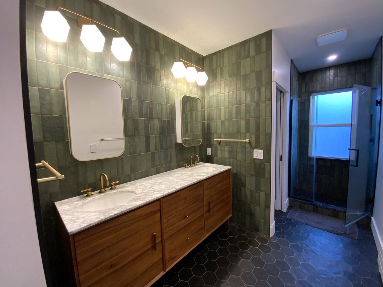 Modern bathroom with dual sinks, marble countertop, hexagonal mirrors, and brass fixtures, green tile walls and dark hexagonal floor tiles, featuring a walk-in shower with frosted glass window.