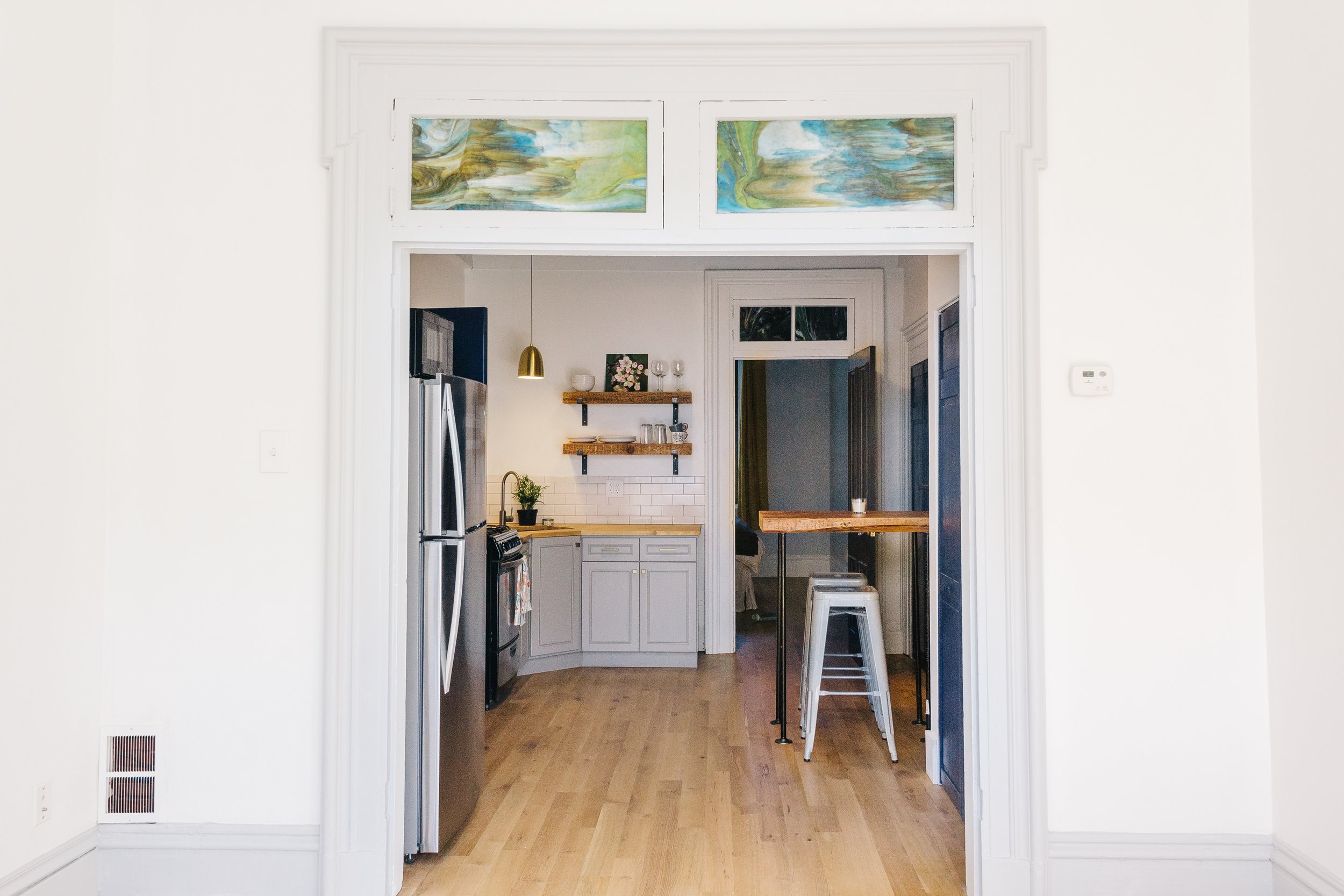 Modern kitchen with stainless steel appliances, open shelving, wooden countertops, pendant lighting, and bar stools. Light-colored walls and flooring, with decorative stained glass panels above the doorway.