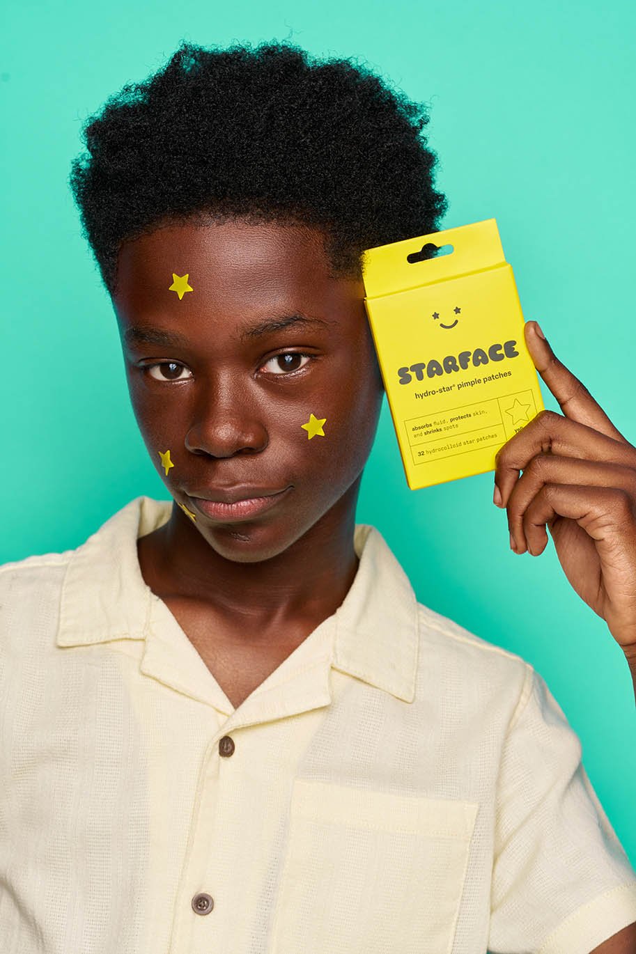 A young person with dark skin and short curly hair, adorned with yellow star-shaped stickers, holds a yellow box labeled 'Starface hydro-star pimple patches' against a teal background.