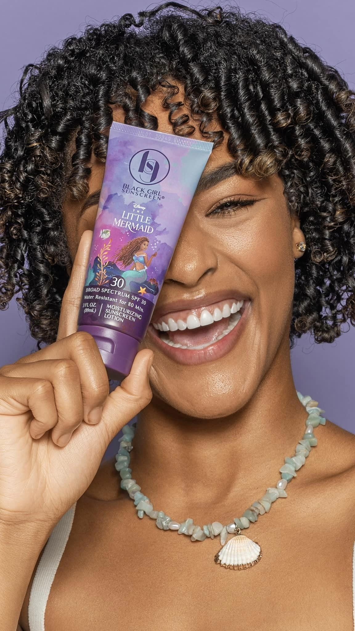 A woman with curly hair smiling and holding a tube of sunscreen labeled 'Black Girl Sunscreen Little Mermaid' with purple background and seashell necklace.