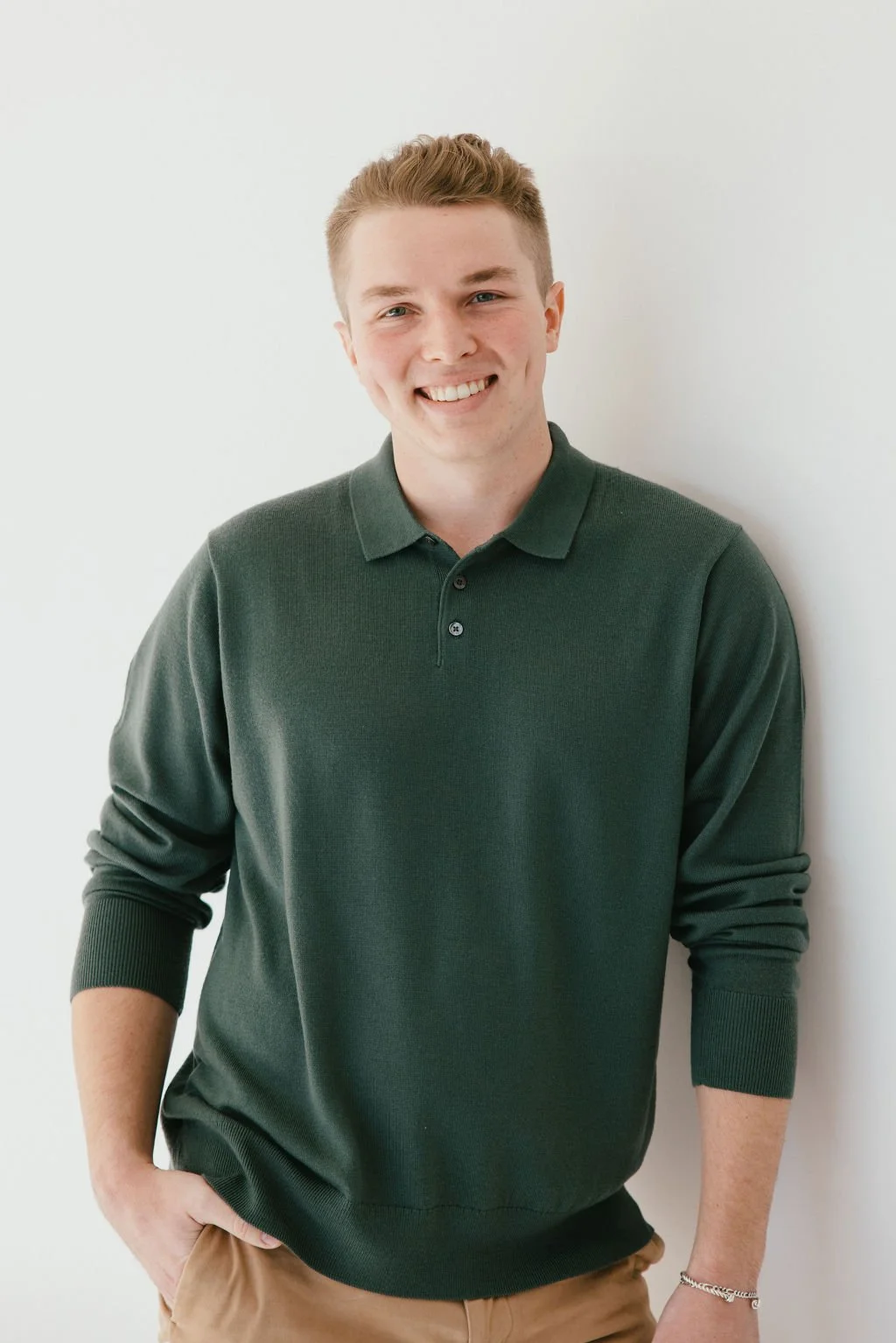 A smiling man with brown hair, wearing a green button up shirt and kaki pants leaning up against a wall