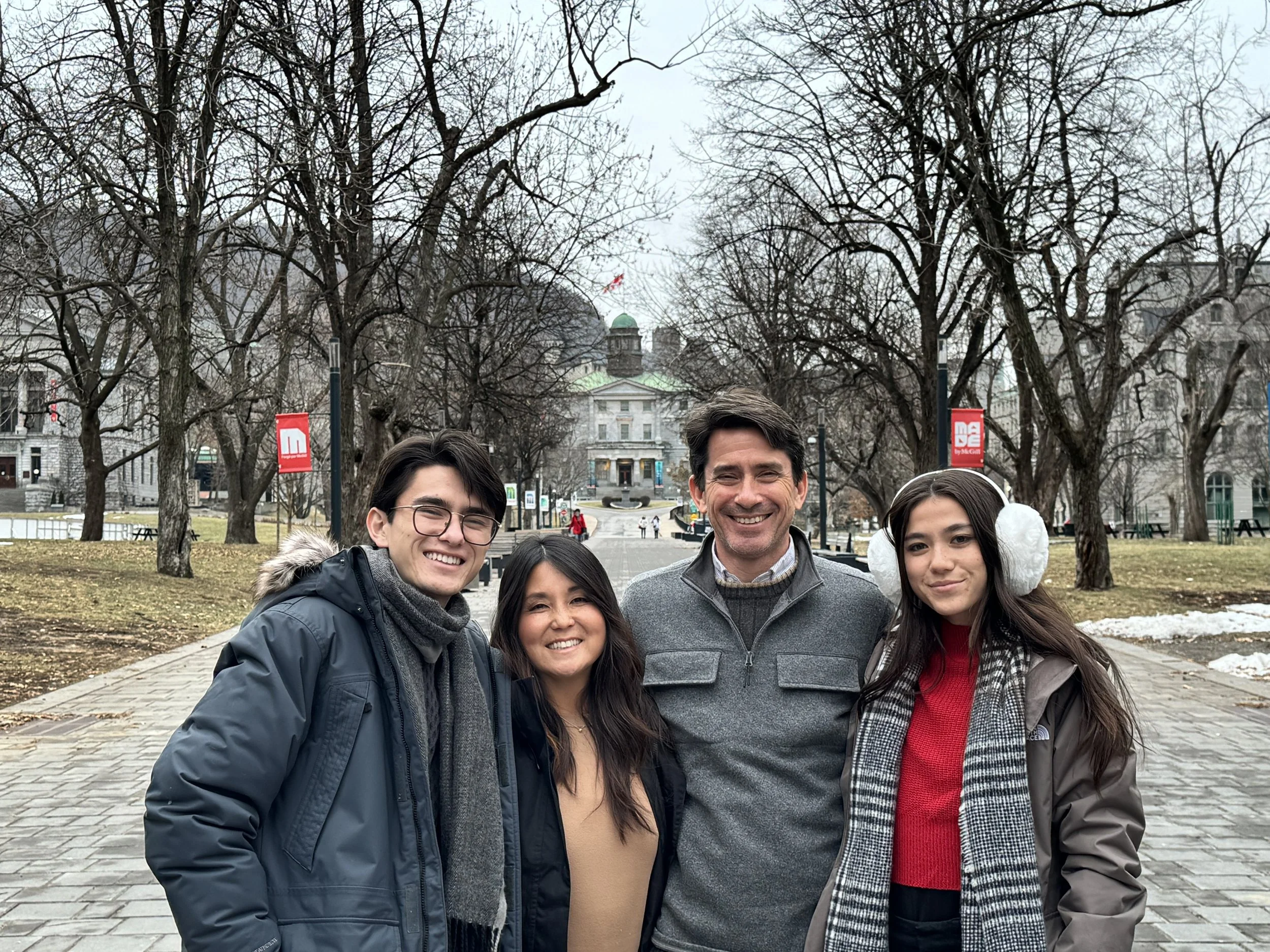 Group of five people posing outdoors in a park with leafless trees and historic buildings in the background.