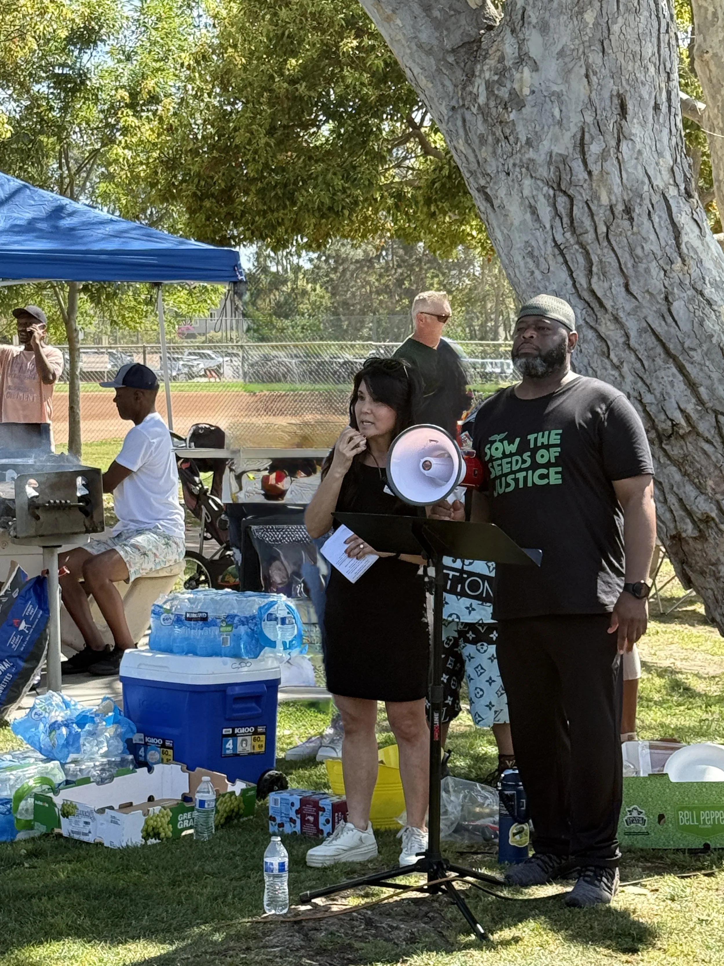 People outdoors at a community gathering or event, standing near tables with food, drinks, and supplies, under a large tree on a sunny day.