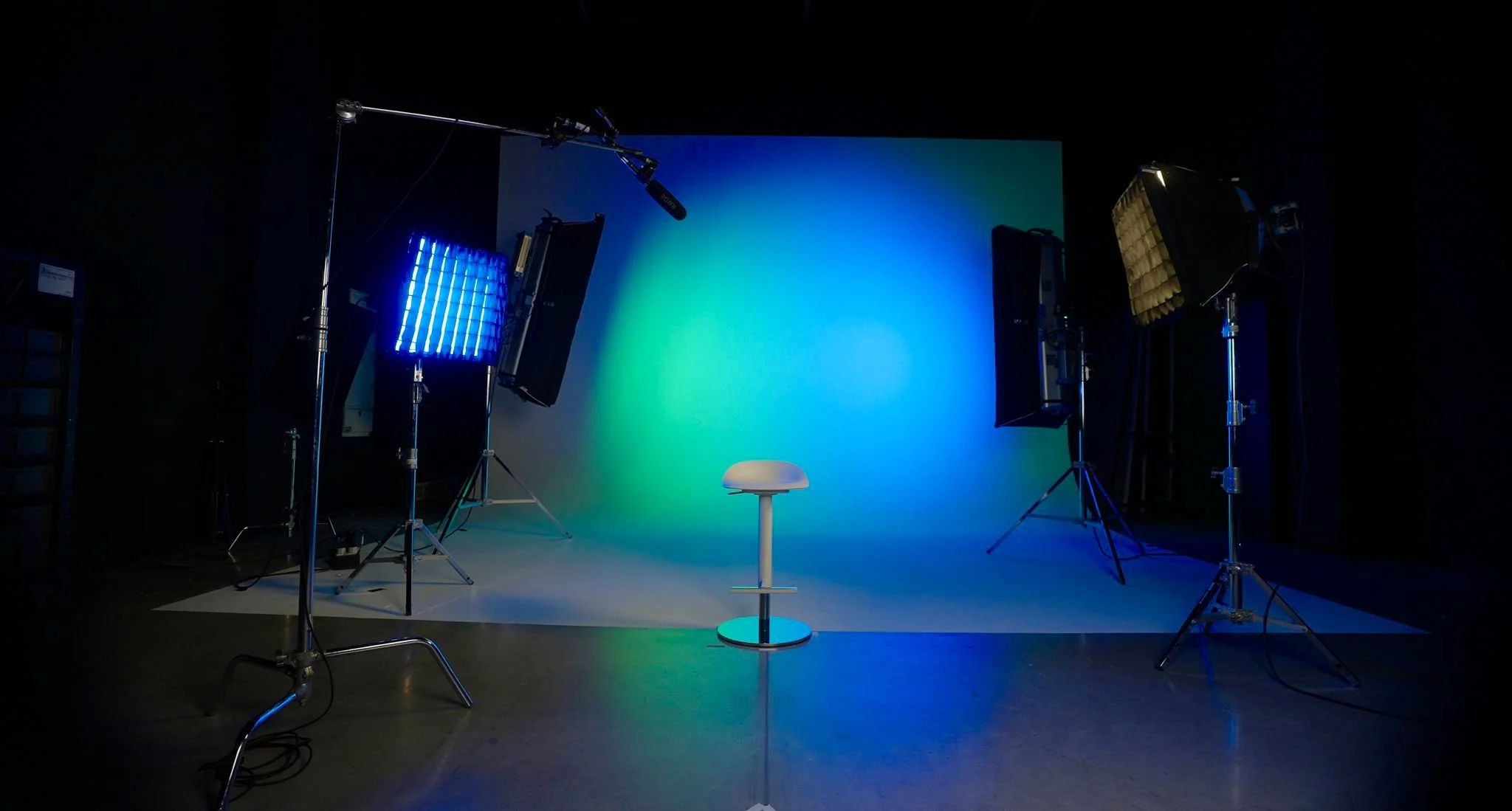 Photo studio with a gray backdrop, professional lighting equipment, a white stool, and colorful lighting effects creating a blue and green glow on the backdrop.