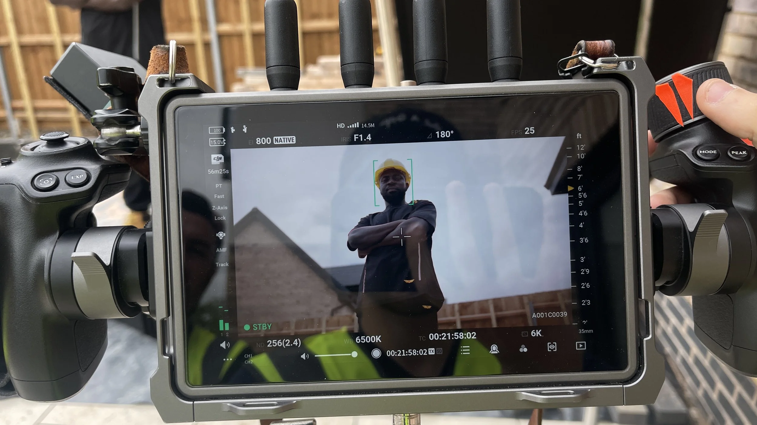 A camera monitor displays a man in a black jacket and yellow hard hat posing outdoors, with construction materials and a wooden structure visible in the background.