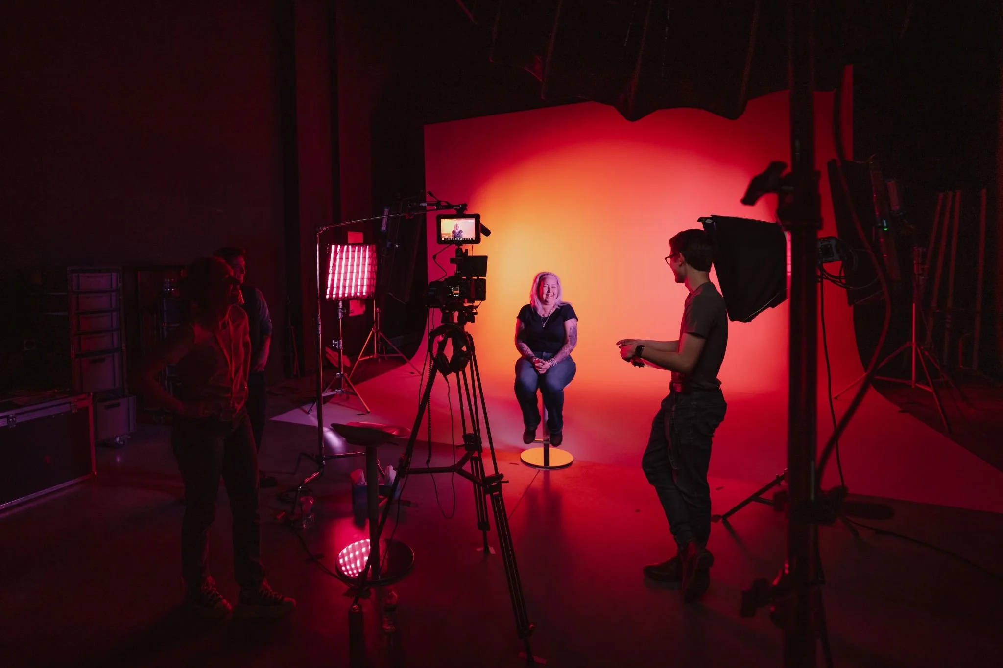 A woman with blonde hair smiling while siting on a stool in a photography studio with a red background, surrounded by camera equipment and crew members.