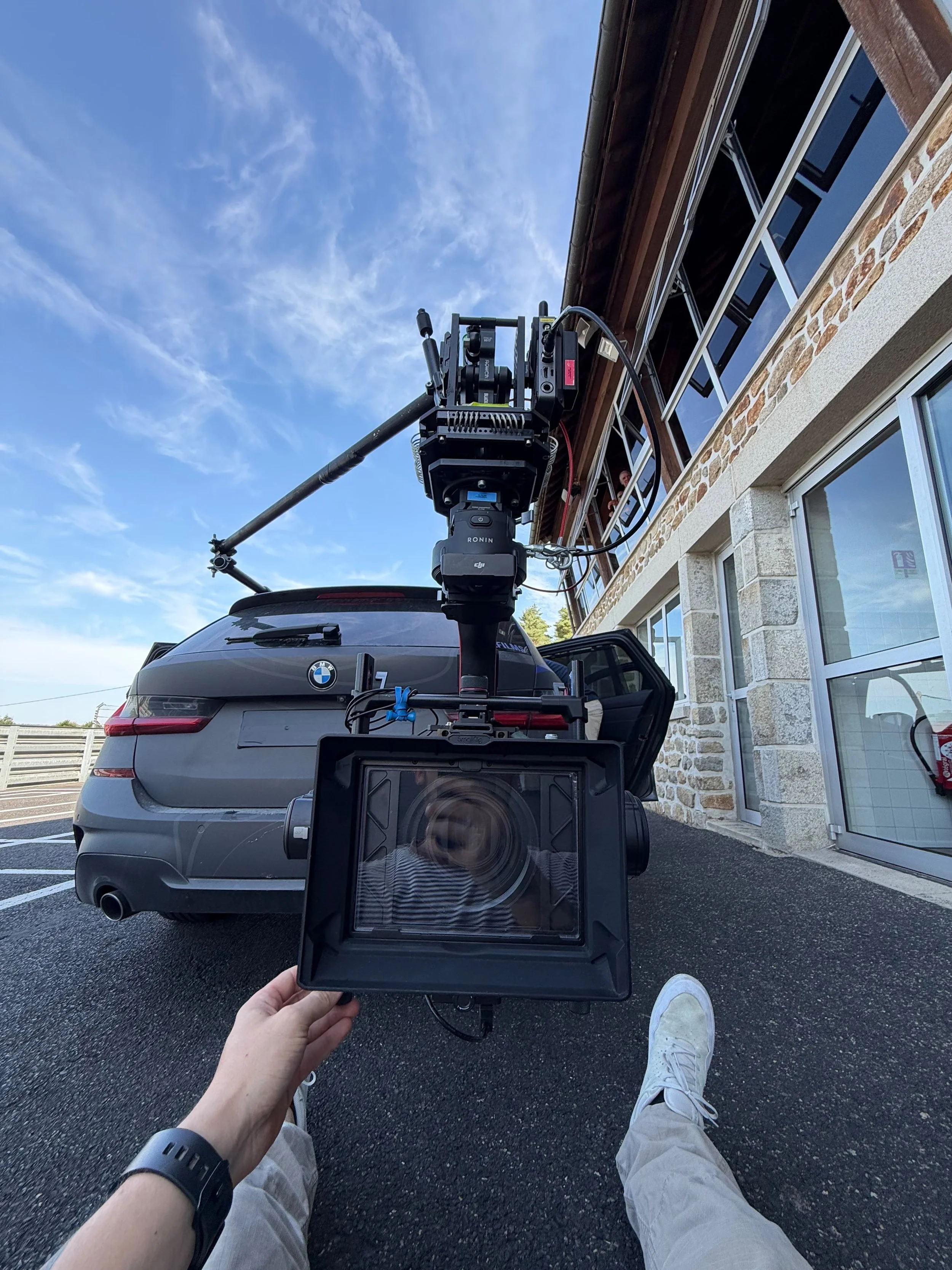Person holding a camera matte box attached to tracking vehicle for filming, with a person’s legs and white sneakers visible, sitting on the ground outside a building with a BMW parked nearby, under a blue sky with some clouds.