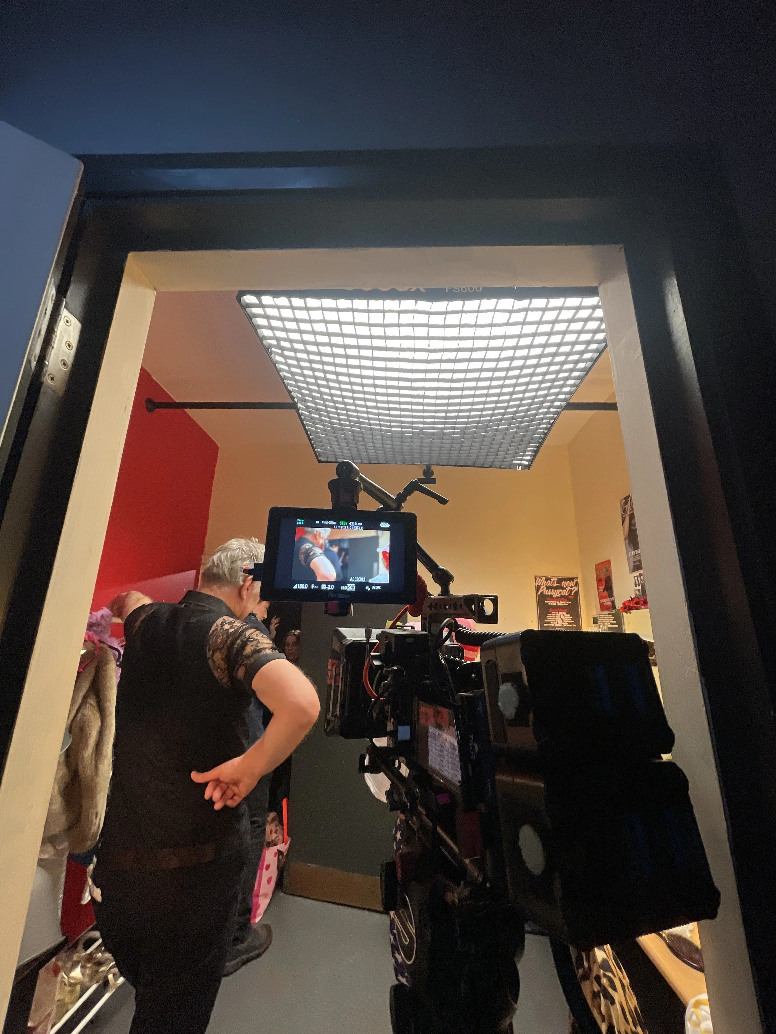 People gathered around a camera setup in a room with yellow and red walls, with a large, bright, square light fixture on the ceiling.