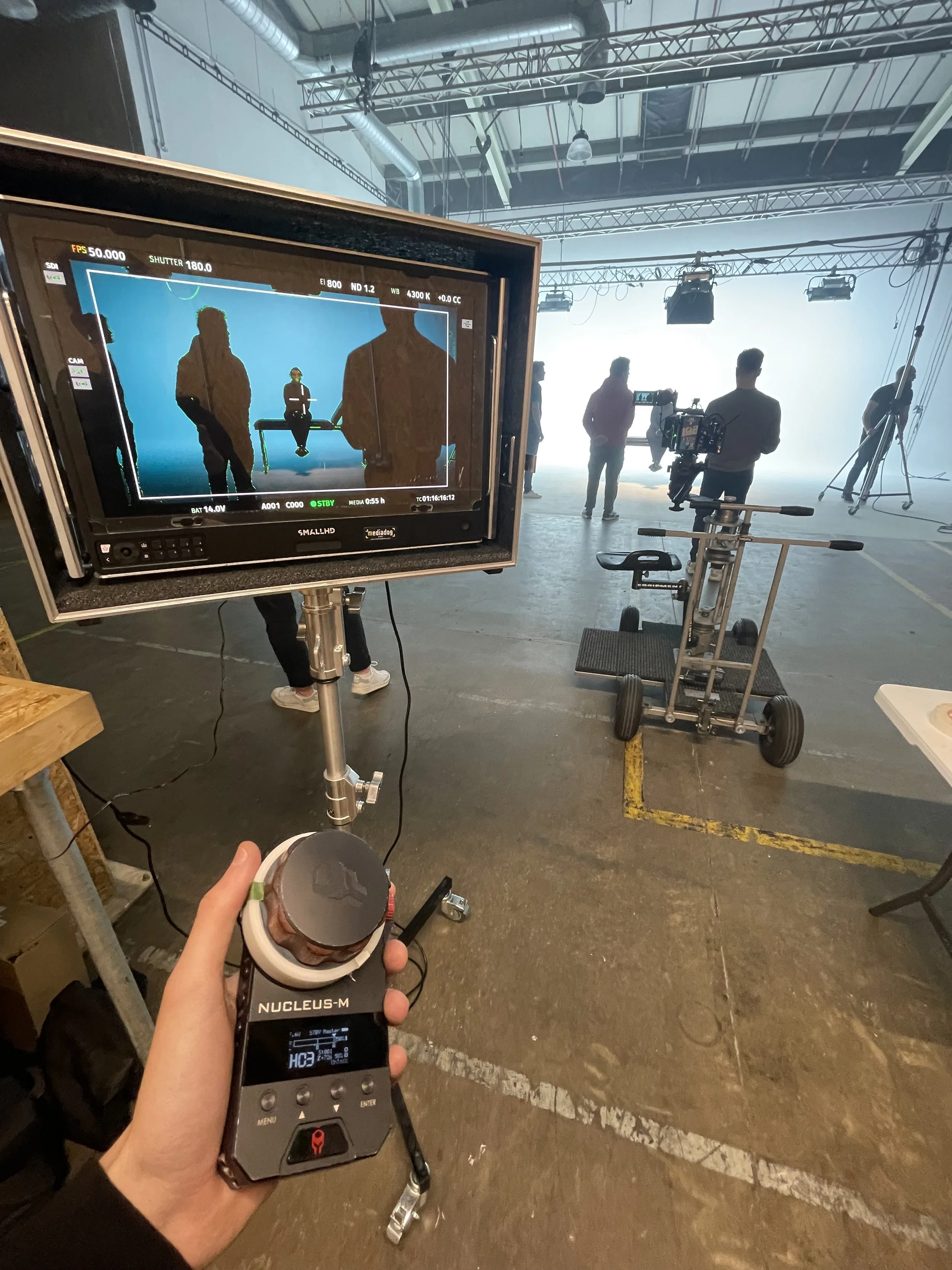 Behind the scenes of a film or photo shoot with a large monitor displaying silhouettes of people, a person holding a measurement device, and group of crew members in a studio with lighting equipment and scaffolding.