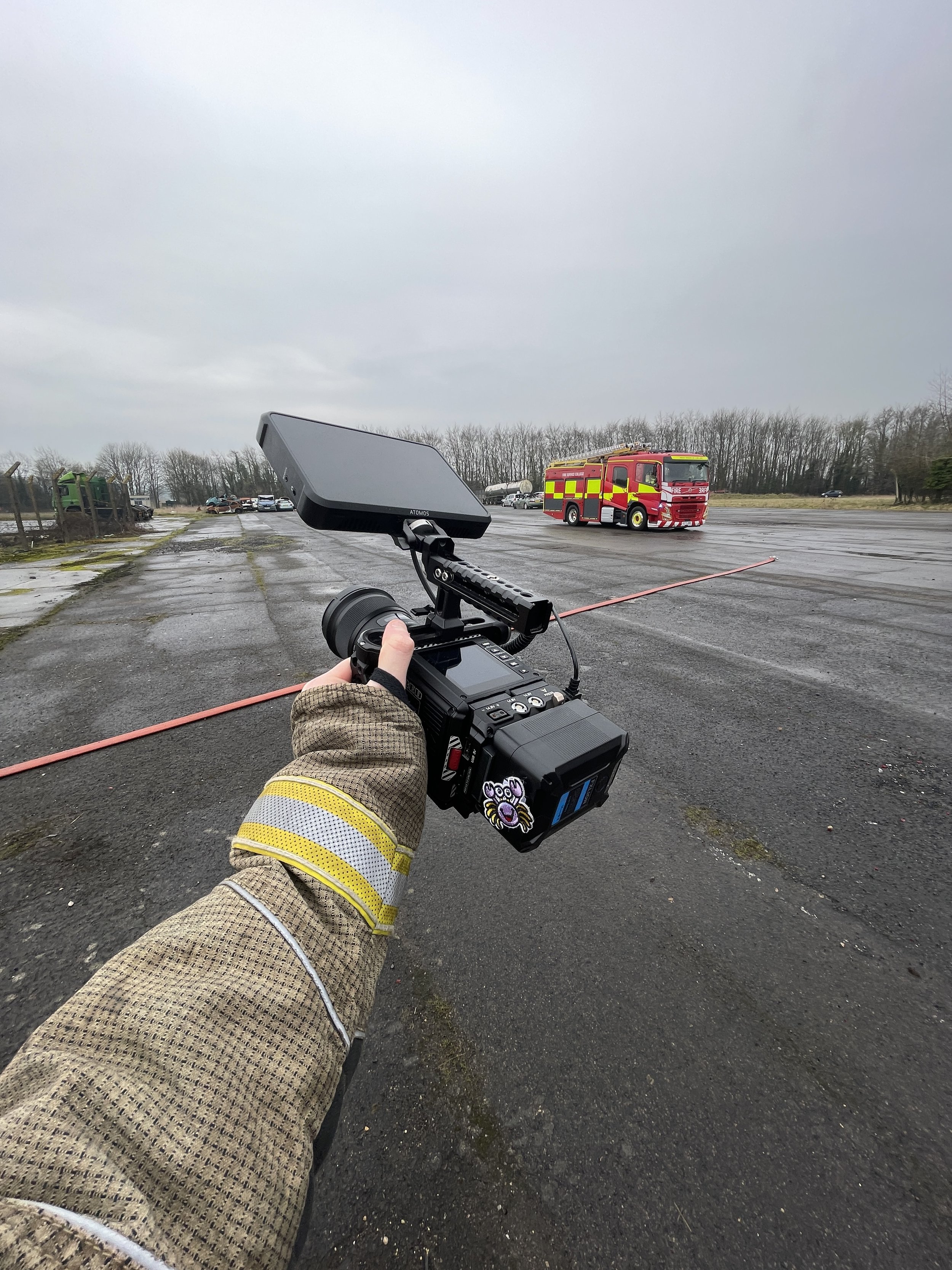 A firefighter holding a camera captures an emergency scene with a red fire truck and several parked cars on an overcast day with a cloudy sky.