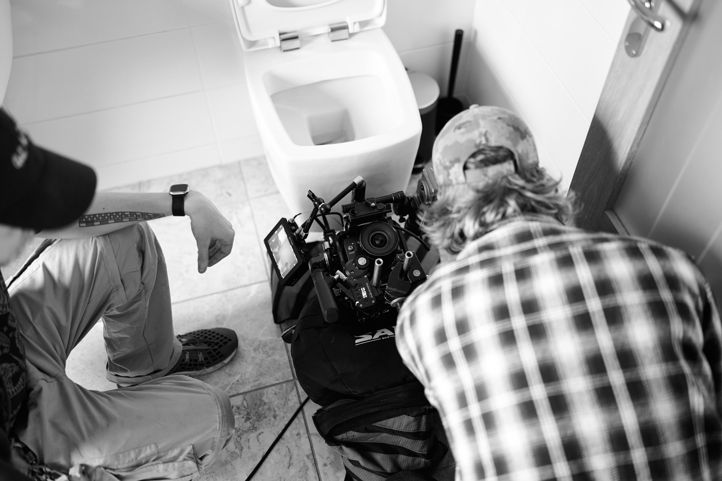 Two crew members filming behind the scenes in a bathroom, one operating a camera on a tripod and the other sitting on the floor nearby.