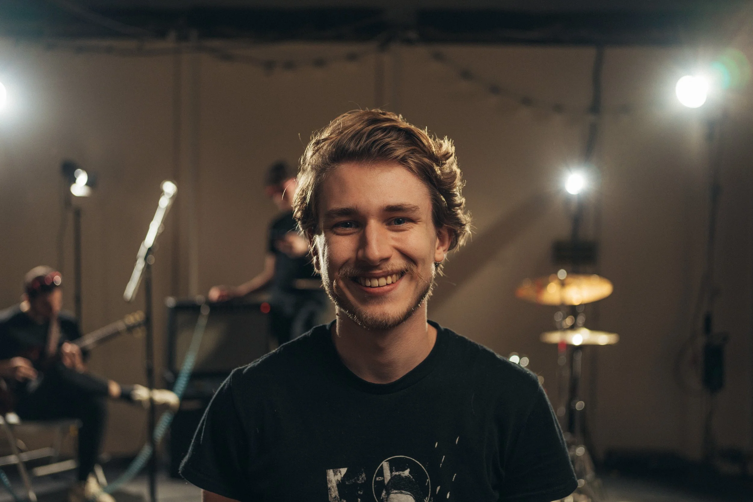 A young man smiling in the foreground with a band and music equipment blurred in the background in a recording studio.