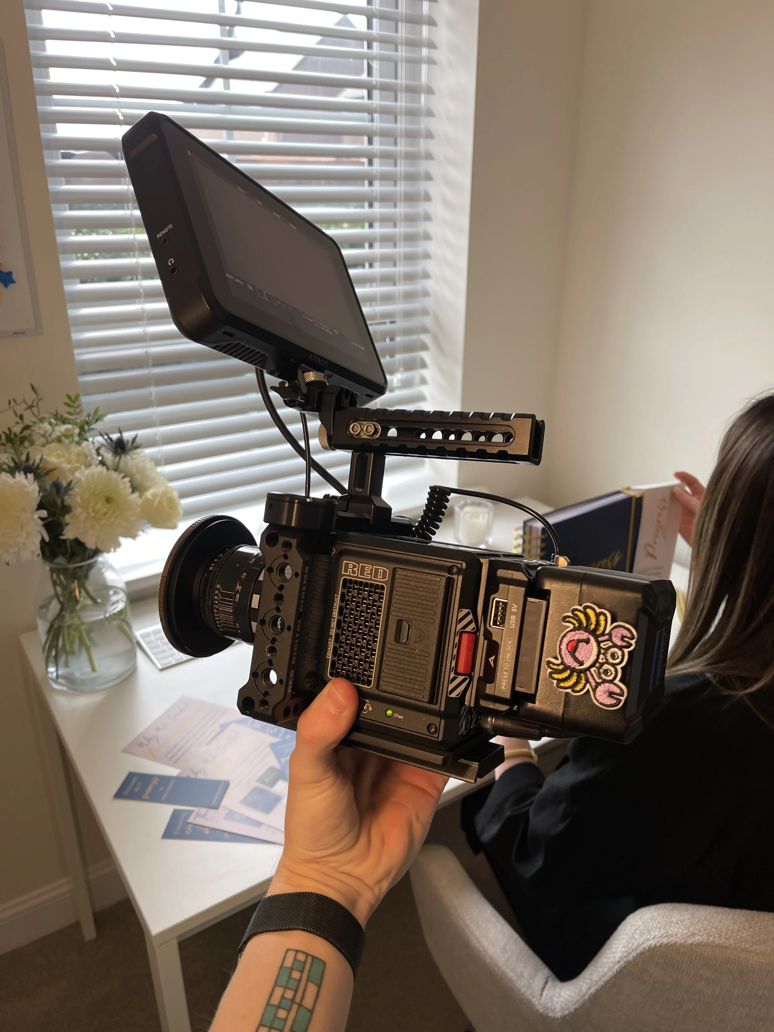 A person holding a professional camera with a sticker on it, pointed at another person who is sitting on a chair with a book. The background shows a window with blinds and a table with brochures and a vase of white flowers.