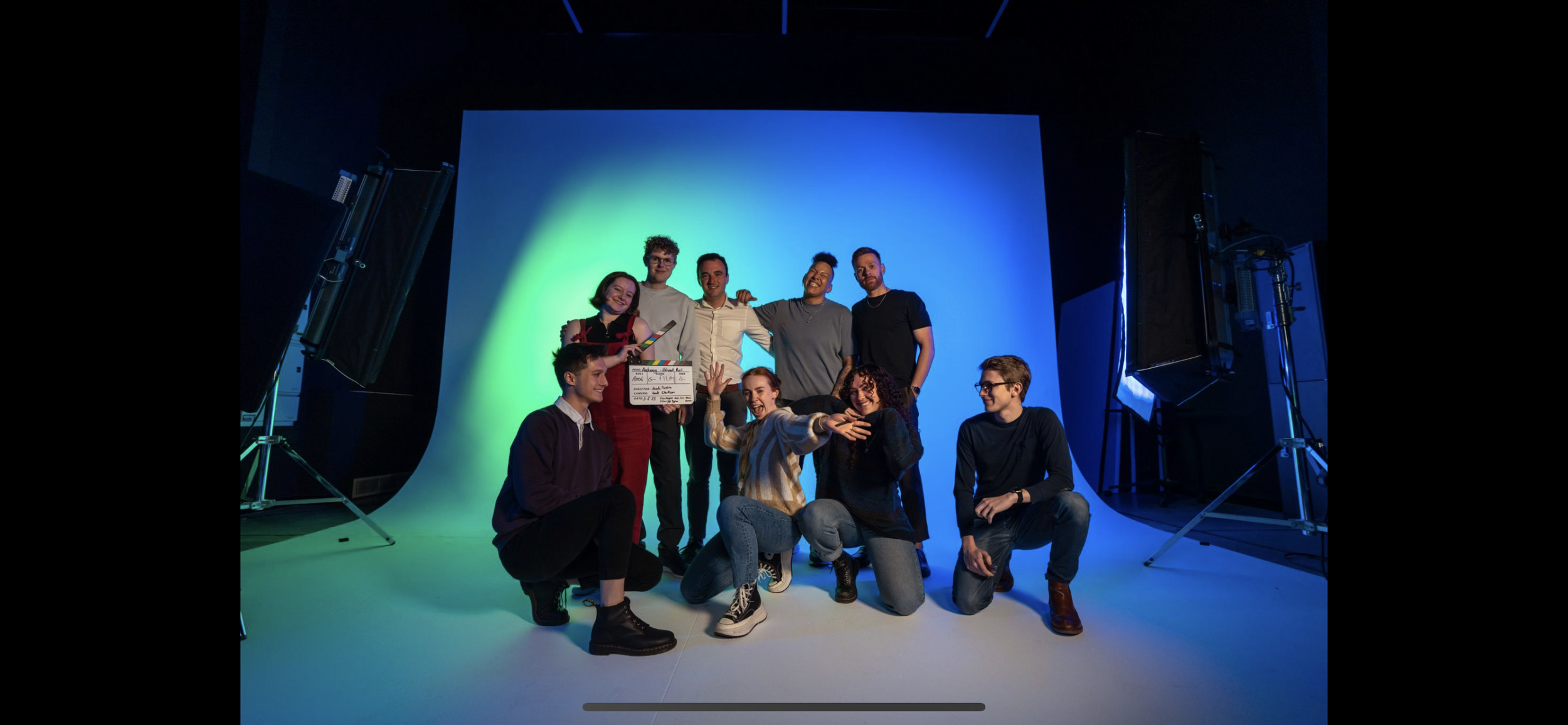A group of nine diverse young people are posing for a photo in a photography studio with a large white backdrop. They are smiling and appear to be celebrating, with some standing and others sitting or crouching. One person is holding a clapperboard. 