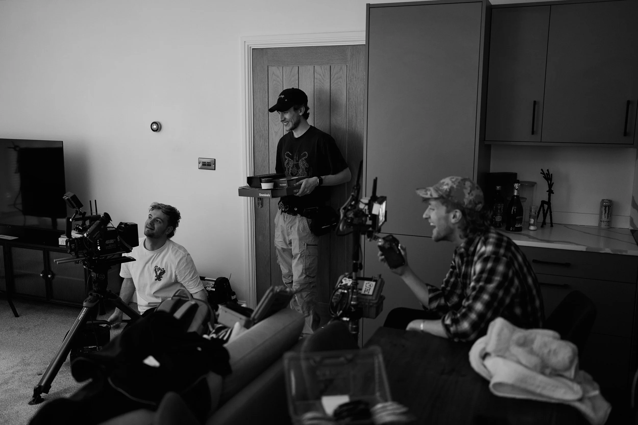Group of young men working with camera equipment and filming inside a room with kitchen cabinets and a wooden door.