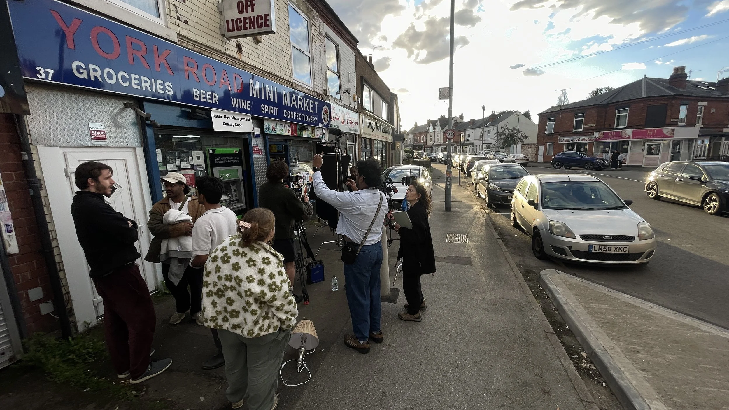 A group of people, including media crew with cameras and microphones, gathered outside a store called York Road Mini Market, with a sign indicating it offers groceries, beer, wine, spirits, and confectionery. Several parked cars line the street, and 