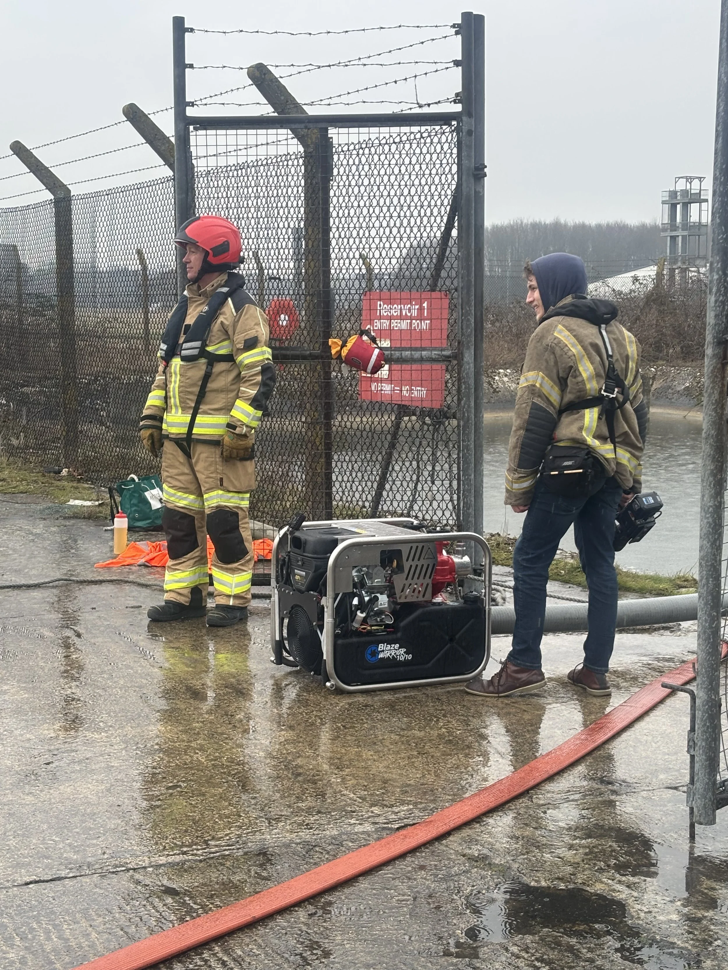 Firefighters and a person with camera gear standing next to a fenced area with barbed wire, a sign that reads 'Receiver 1 Entry Permit Point,' and firefighting equipment on a wet ground with water and hoses.