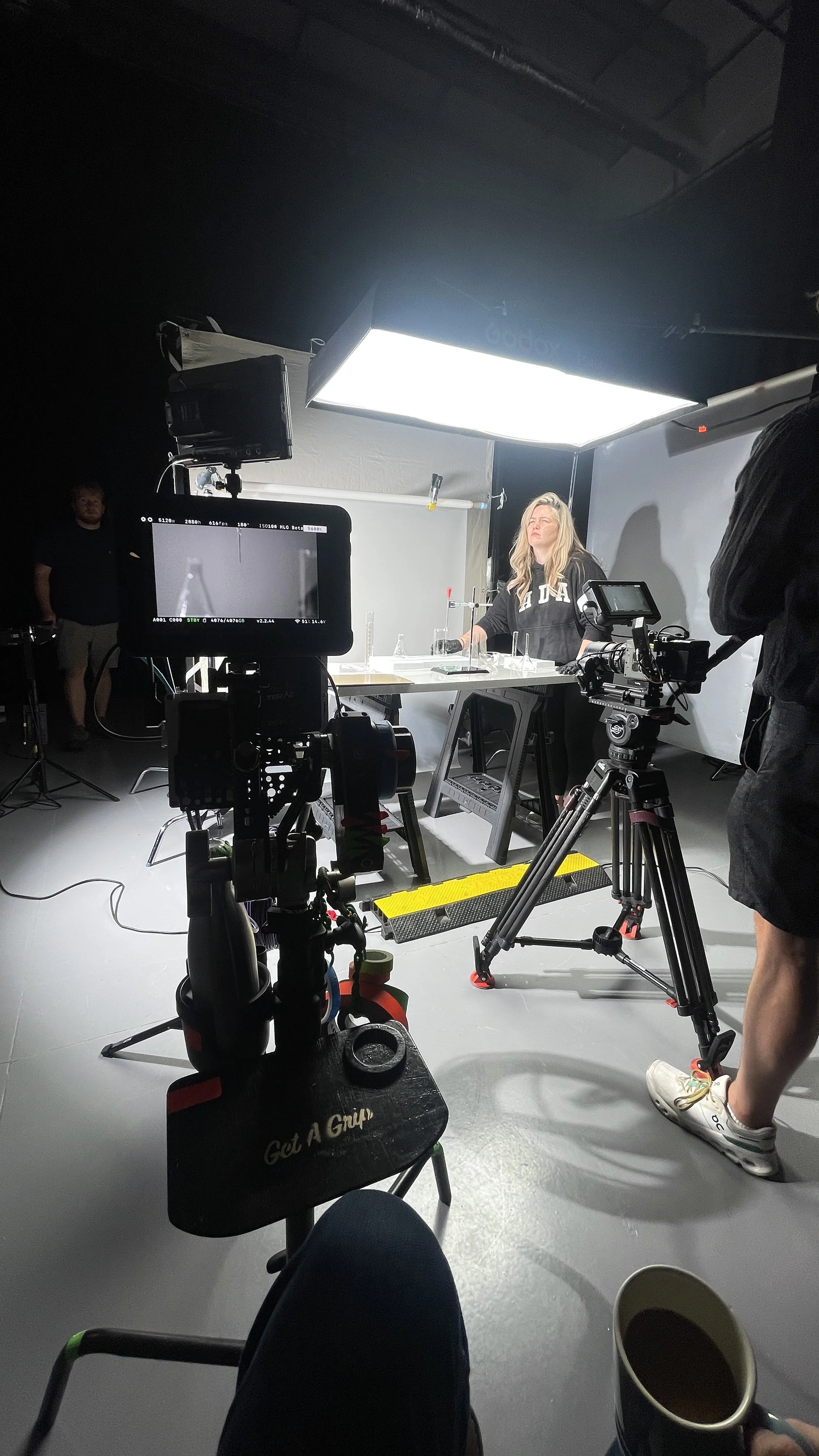A woman in a black hoodie with white and beige writing on it stands behind a table with laboratory glassware in a studio. There are multiple cameras and lighting equipment around her, a crew member on the side, and a person holding a cup of coffee in