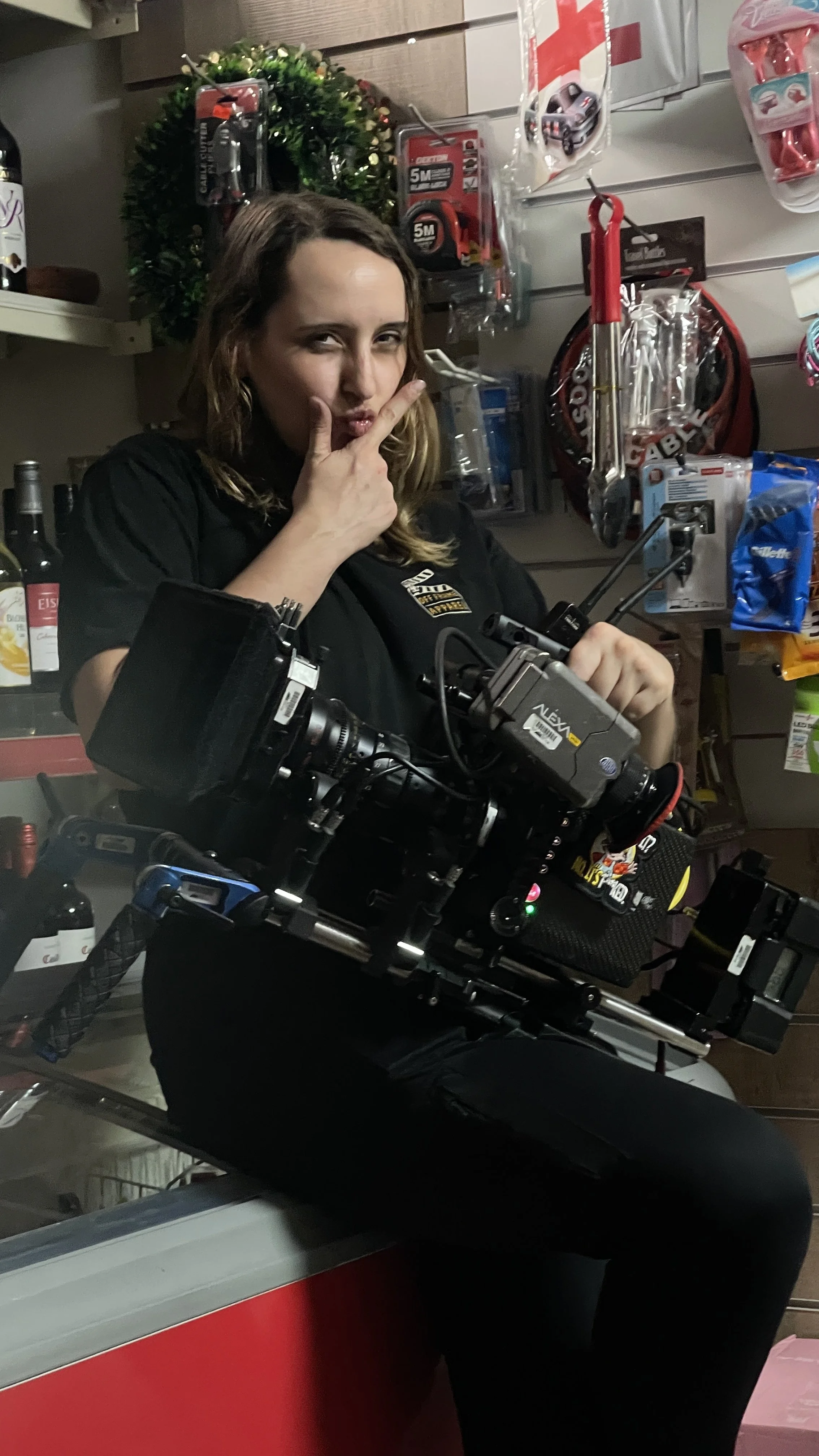 Woman with shoulder-length hair in a store posing with her hand on her chin and a finger pointing upward. She is standing behind a camera setup with various equipment. The background has shelves with tools, accessories, and a decorative wreath.
