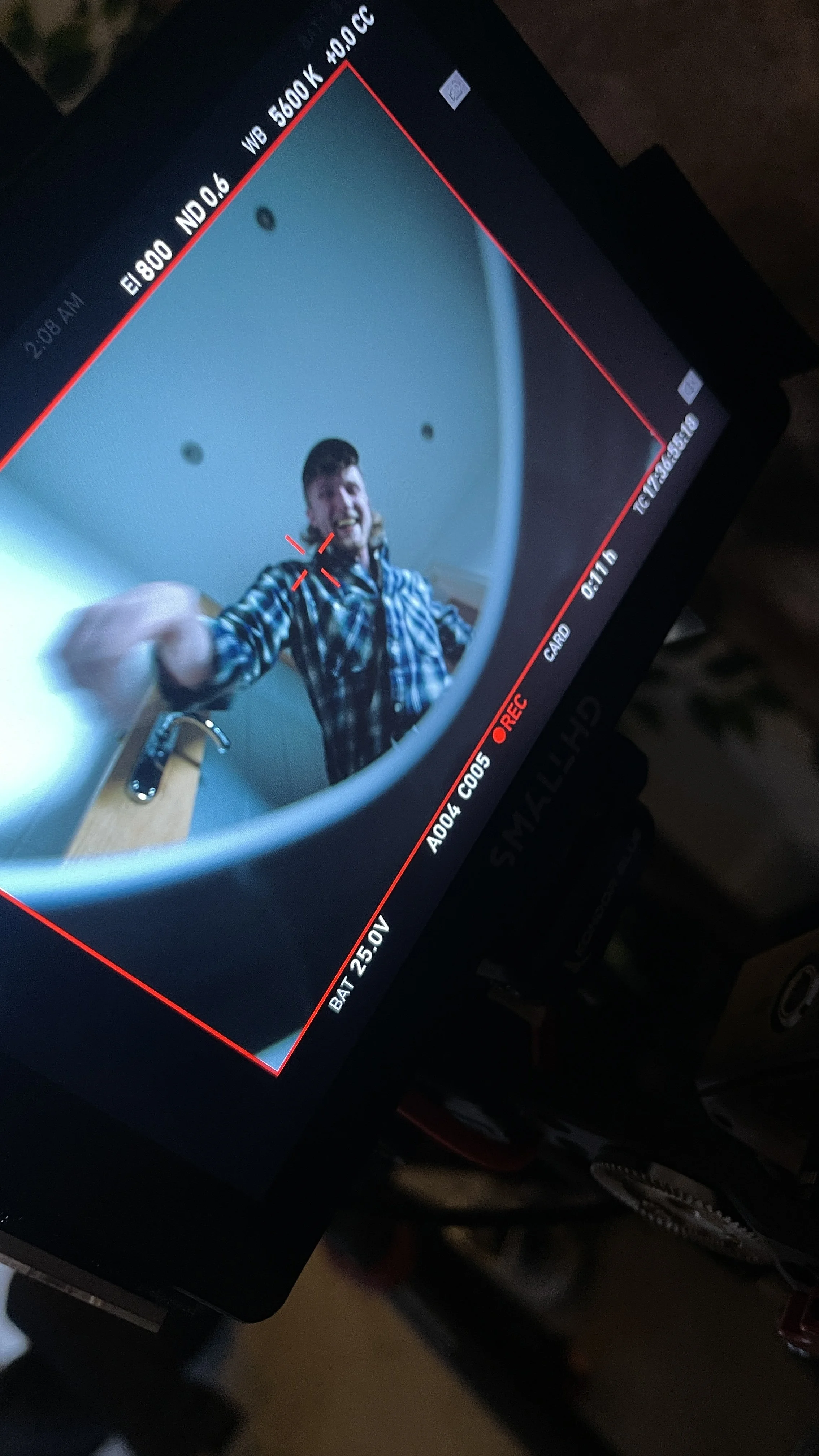 Young man smiling and pointing at the camera, taking a selfie through a fisheye lens on a camera monitor, with a bathroom sink and faucet visible in the background.
