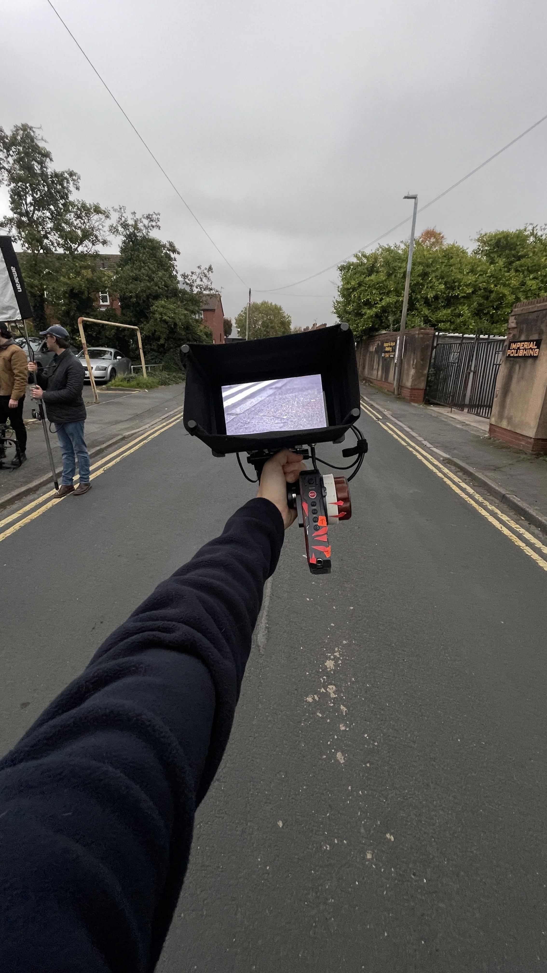 First-person view of a person holding a camera on a gimbal stabilizer, pointed at a street scene with a cloudy sky, trees, and a fence with a sign reading 'Imperial Polishing'; two people are standing on the sidewalk to the left, one holding a microp