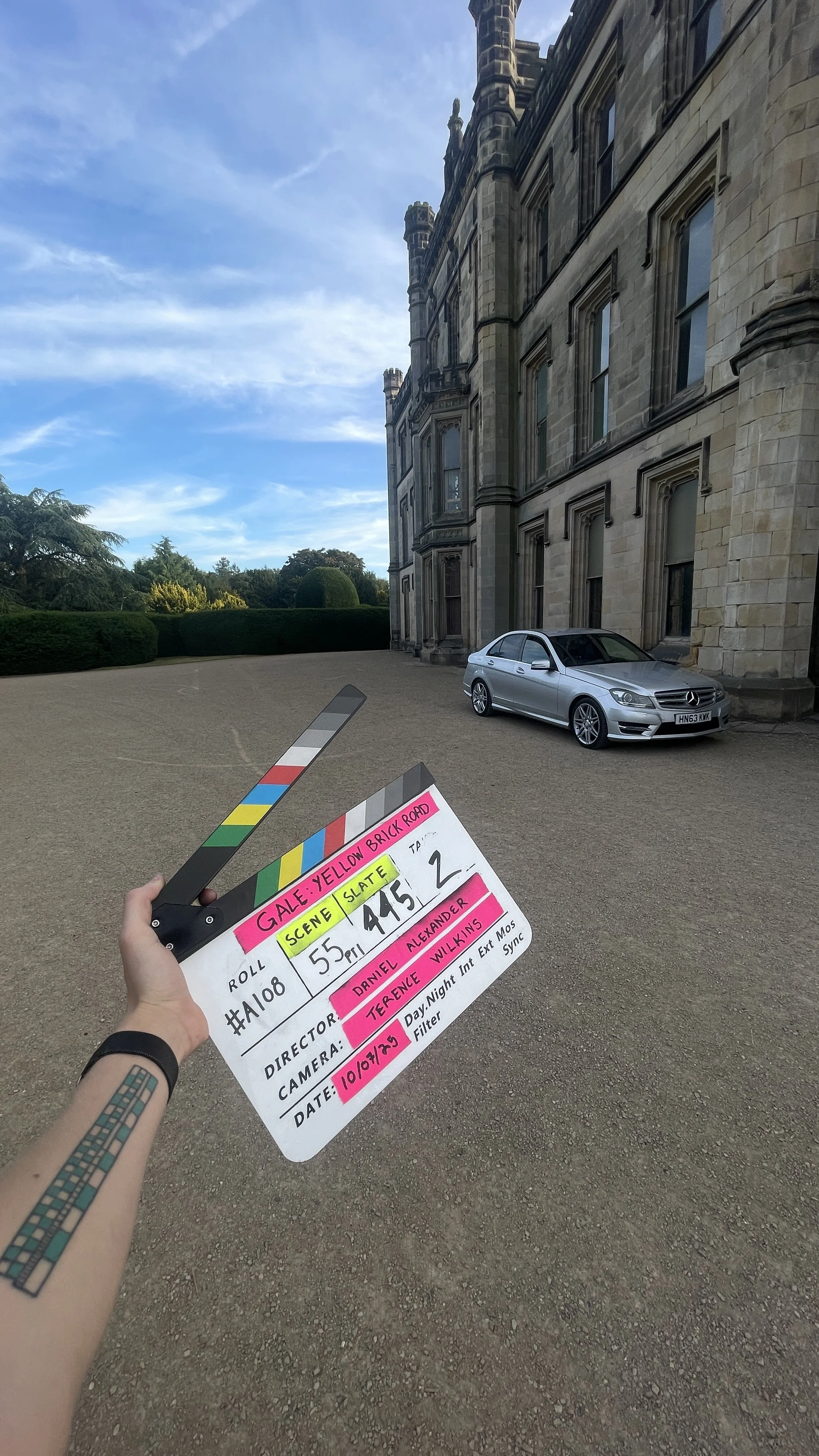 A film slate held by a person's hand in front of a large stone building with a silver car parked nearby and trees in the background.
