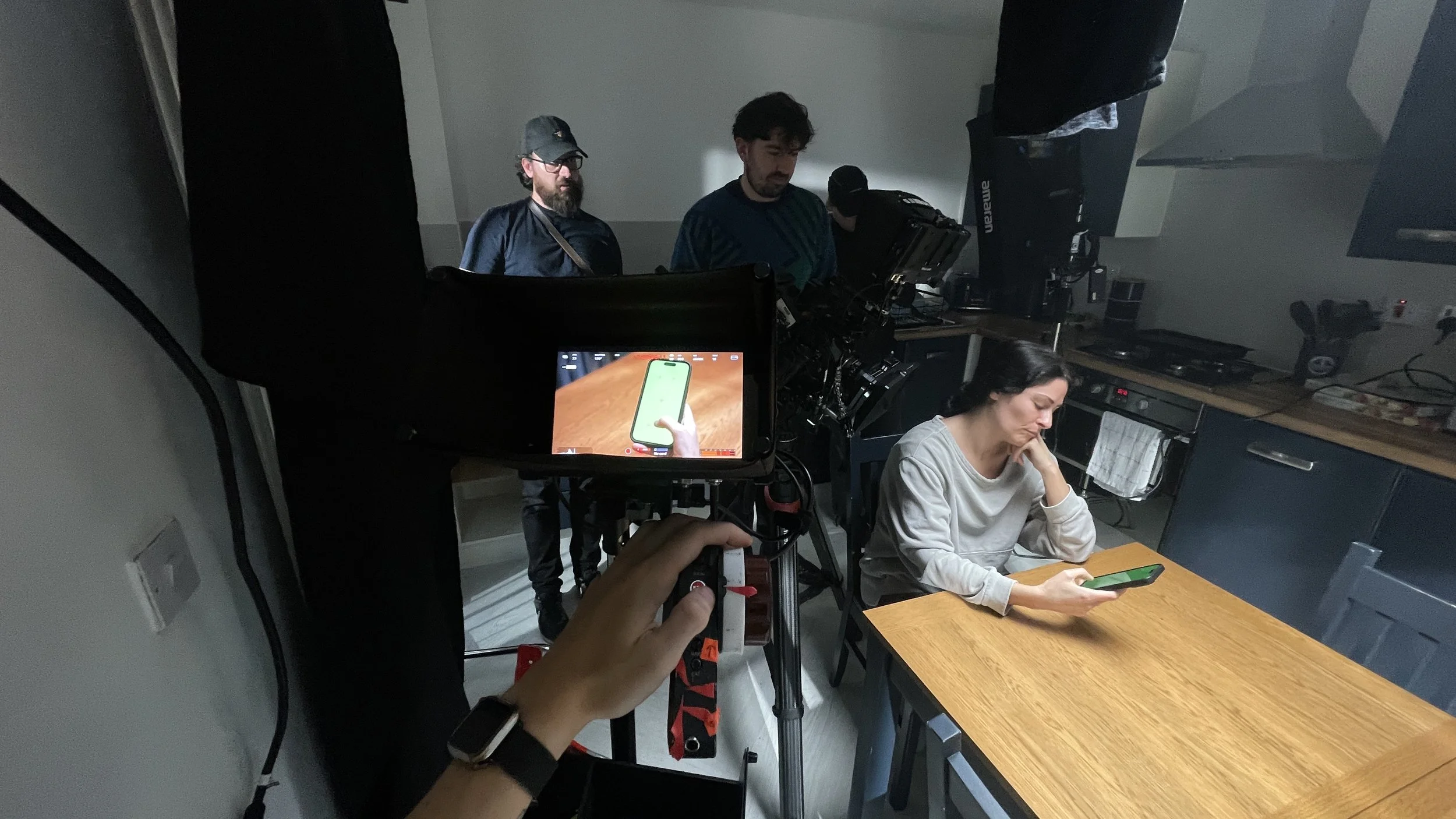 People working behind the scenes of a video or photo shoot in a kitchen. One woman with dark hair, wearing a light gray sweatshirt, seated at a wooden table, looking at her smartphone. Three men standing behind her, observing or operating equipment, 
