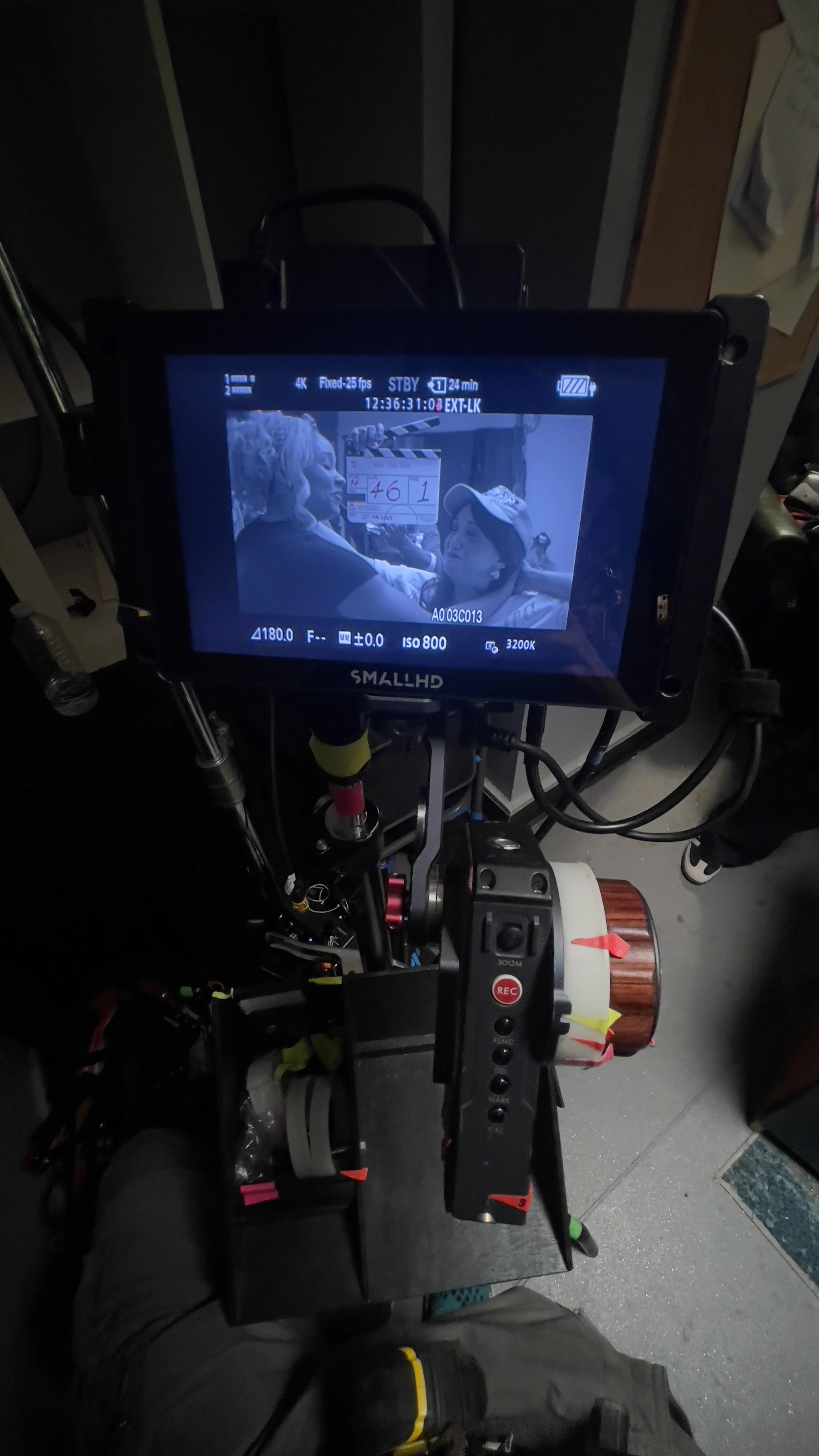 View of a camera monitor on a film set showing two women, one with textured platinum blonde hair and makeup, and the other wearing a cap, engaged in a close moment. The camera setup and equipment are visible in the foreground.