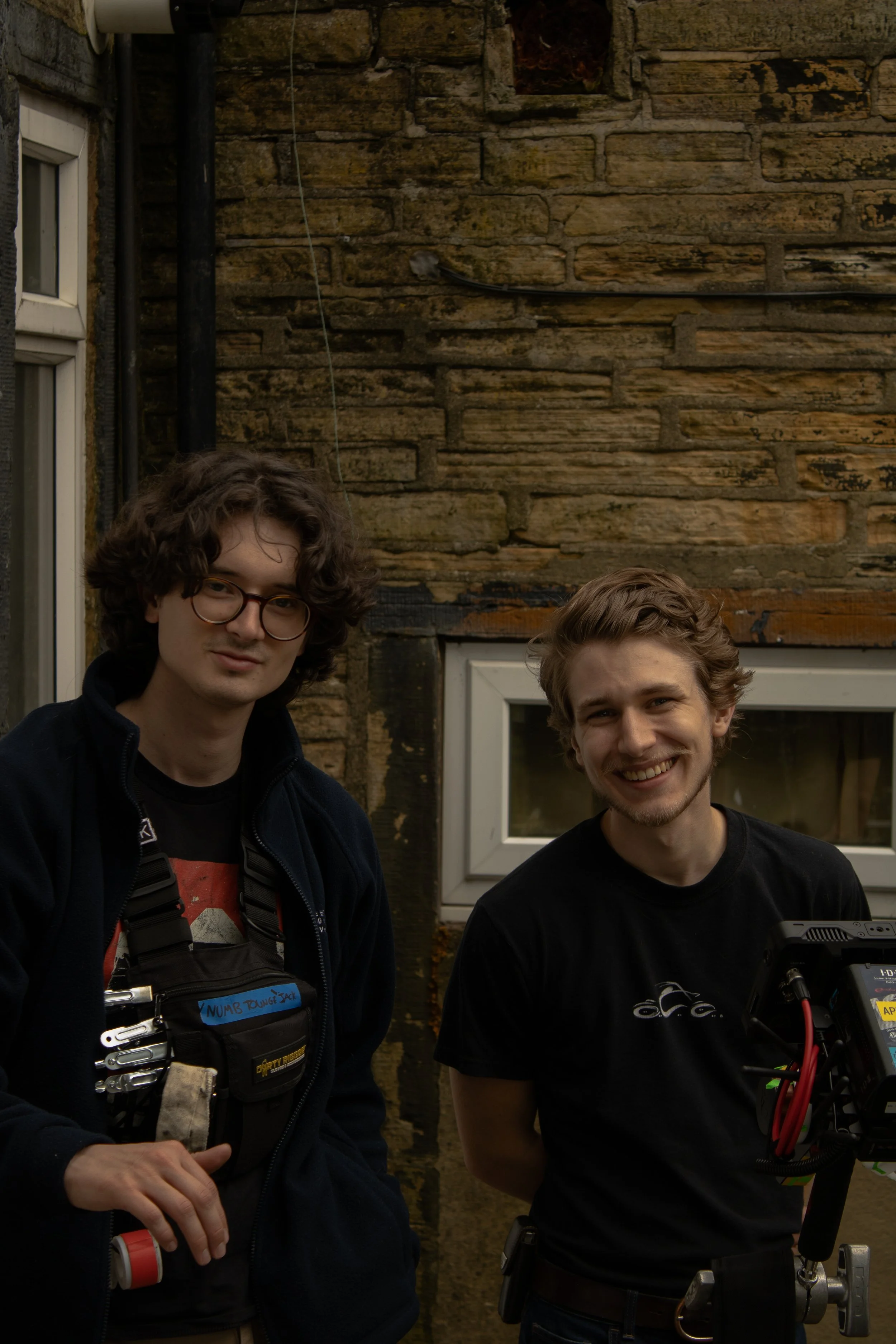Two young men smiling and standing outdoors in front of a brick wall. The person on the left has curly hair, glasses, and a work vest, while the person on the right has wavy hair and is holding camera equipment.